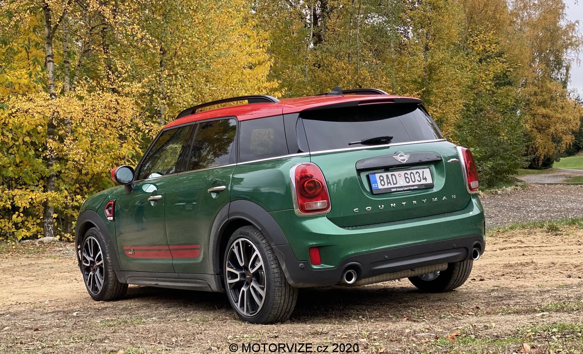 Trojštvrťový zadný pohľad na Mini Cooper Countryman John Cooper Works 2020 s dvojfarebným tmavozeleným a čiernym lakom, zrkadlovými krytkami v červenej farbe, viditeľným emblémom Mini a nápisom 'Countryman', charakteristickými zadnými svetlami, výrazným zadným nárazníkom s parkovacími senzormi, zadným oknom s odmlžovačom, dvojitými výfukovými rúrami, zadným stieračom, päťramennými zliatinovými kolesami, zaparkovaným na poľnej ceste s stromami a jesennými listami v pozadí, zachyteným z miernem vysokého uhla.