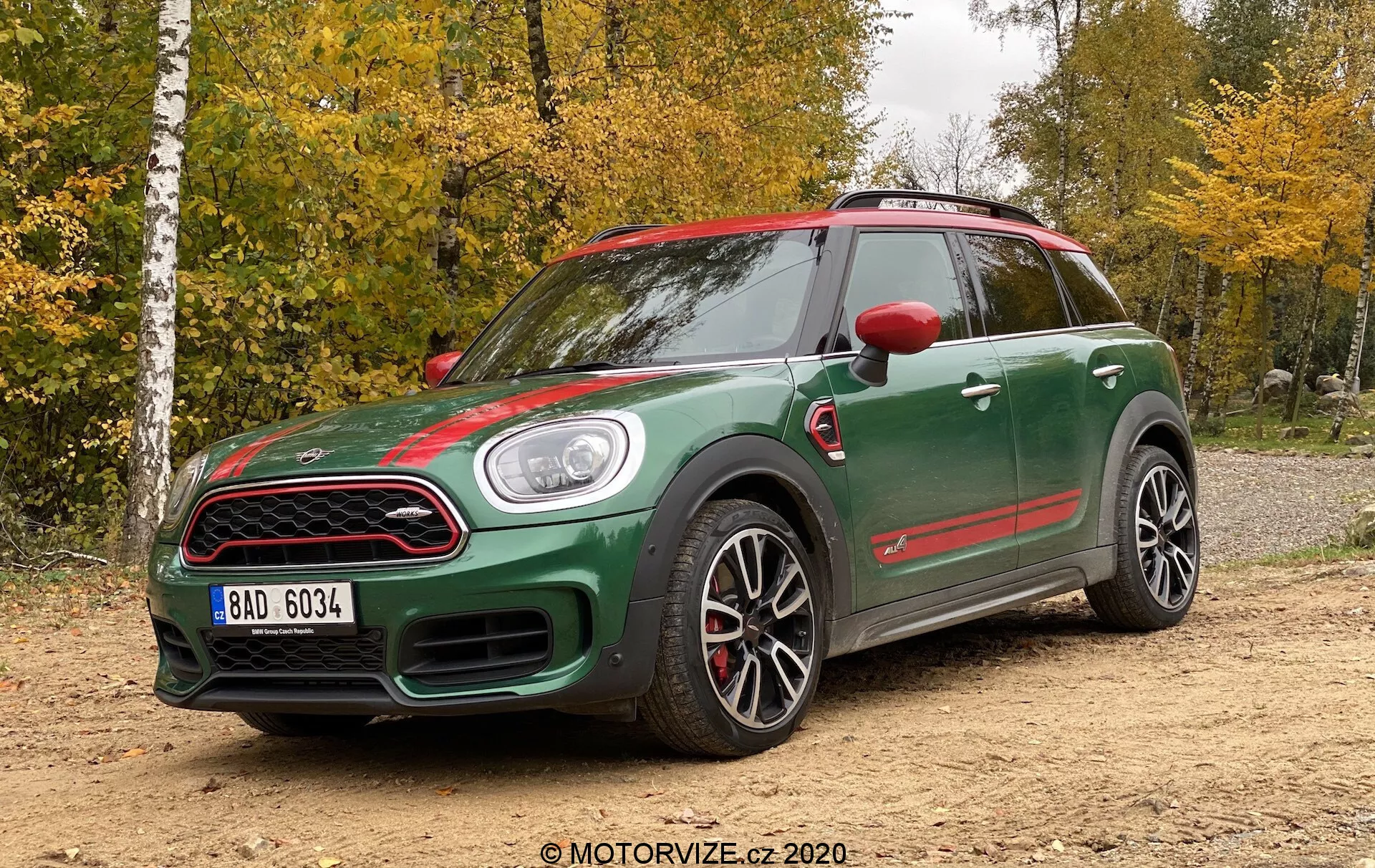 Pohľad z troch štvrtín spredu na Mini Countryman John Cooper Works z roku 2020, ktorý ukazuje športový dizajn s dvojtónovou tmavozelenou karosériou a červenými akcentmi na bočných zrkadlách, prednej mriežke a bočných sukniach. Funkcie zahŕňajú výraznú kapotu, okrúhle svetlá, prednú mriežku s medovou sieťovinou, veľké športové zliatinové kolesá s viacodpružkovým dizajnom, červené brzdové strmene, výrazné bočné prahy a agresívny postoj. Vozidlo je umiestnené na neosypanom povrchu s lesným prostredím v pozadí, za prirodzeného svetla pri zamračenom dni.