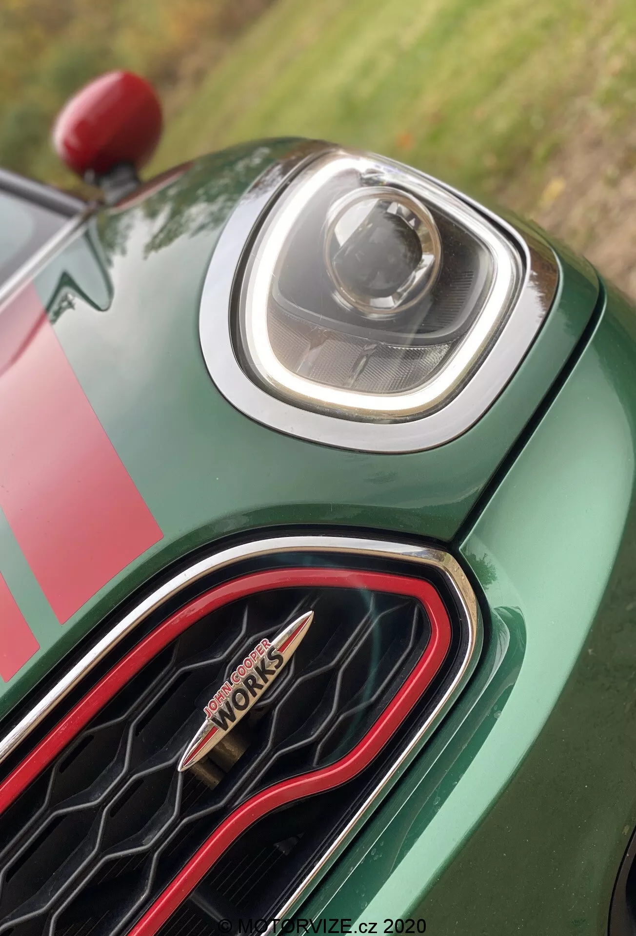 Zblízka pohľad na prednú časť vozidla Mini Countryman John Cooper Works 2020, ktoré zobrazuje pokročilý LED svetlomet a jedinečnú šesťuholníkovú masku s červeným obrysom a odznakom 'John Cooper Works'. Karoséria vozidla má bohatý zelený lak so závodným pruhovým motívom. Foto: PRODRIVE.CZ, 2020.