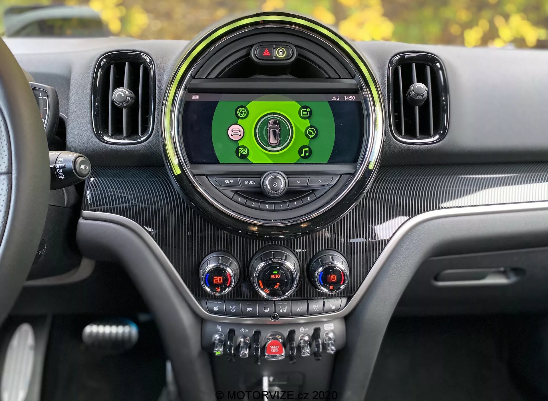 Interiér prístrojovej dosky Mini Countryman John Cooper Works 2020 z pohľadu vodiča. Stredová konzola s veľkým okrúhlym displejom infotainmentu zobrazujúcim farebné ikony pre médiá, telefónne pripojenie, navigáciu a nastavenia vozidla. Orámovaná štýlovým okrúhlym rámom a po stranách sú ďalšie vetracie mriežky. Pod displejom sú analógové ciferníky s chrómovými akcentmi na ovládanie klimatizácie a spínače na funkcie ako vyhrievanie sedadiel, odmrazovanie, recirkulácia vzduchu a jazdné režimy. Textúra podobná karbónu a kovové akcenty poskytujú moderný a luxusný interiérový dizajn s viditeľným digitálnym prístrojovým panelom indikujúcim, že vozidlo je zapnuté.