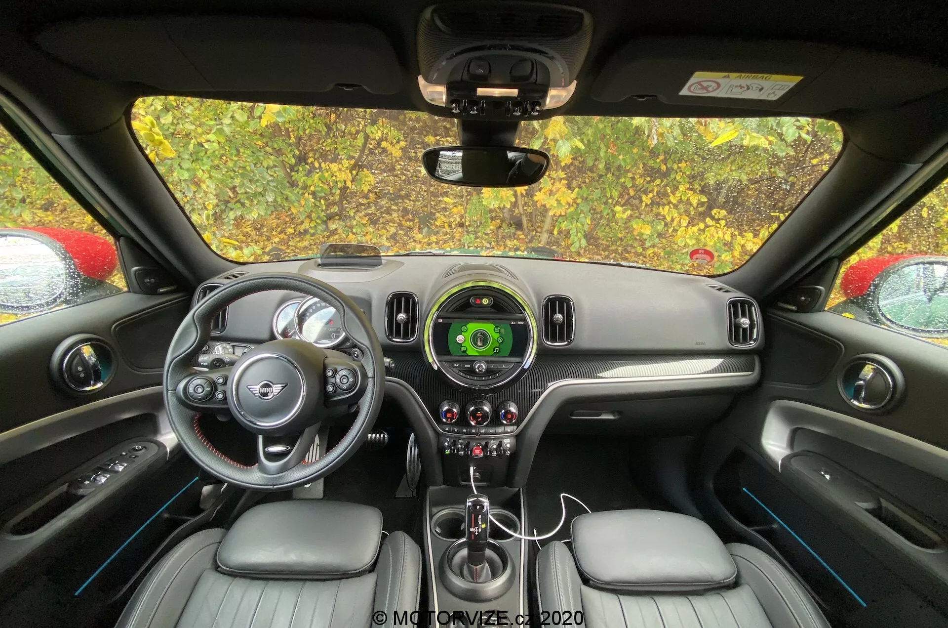 Interiér Mini Countryman 2020 John Cooper Works z perspektívy zadného sedadla, sústredený na palubnú dosku a ovládaciu oblasť vodiča. Volant so striebornými akcentmi a logom značky, centrálny rýchlomer s digitálnym displejom, elegantný dizajn palubnej dosky, radiaca páka, ovládače klímy, kožené sedadlá a spätné zrkadlo sú viditeľné.