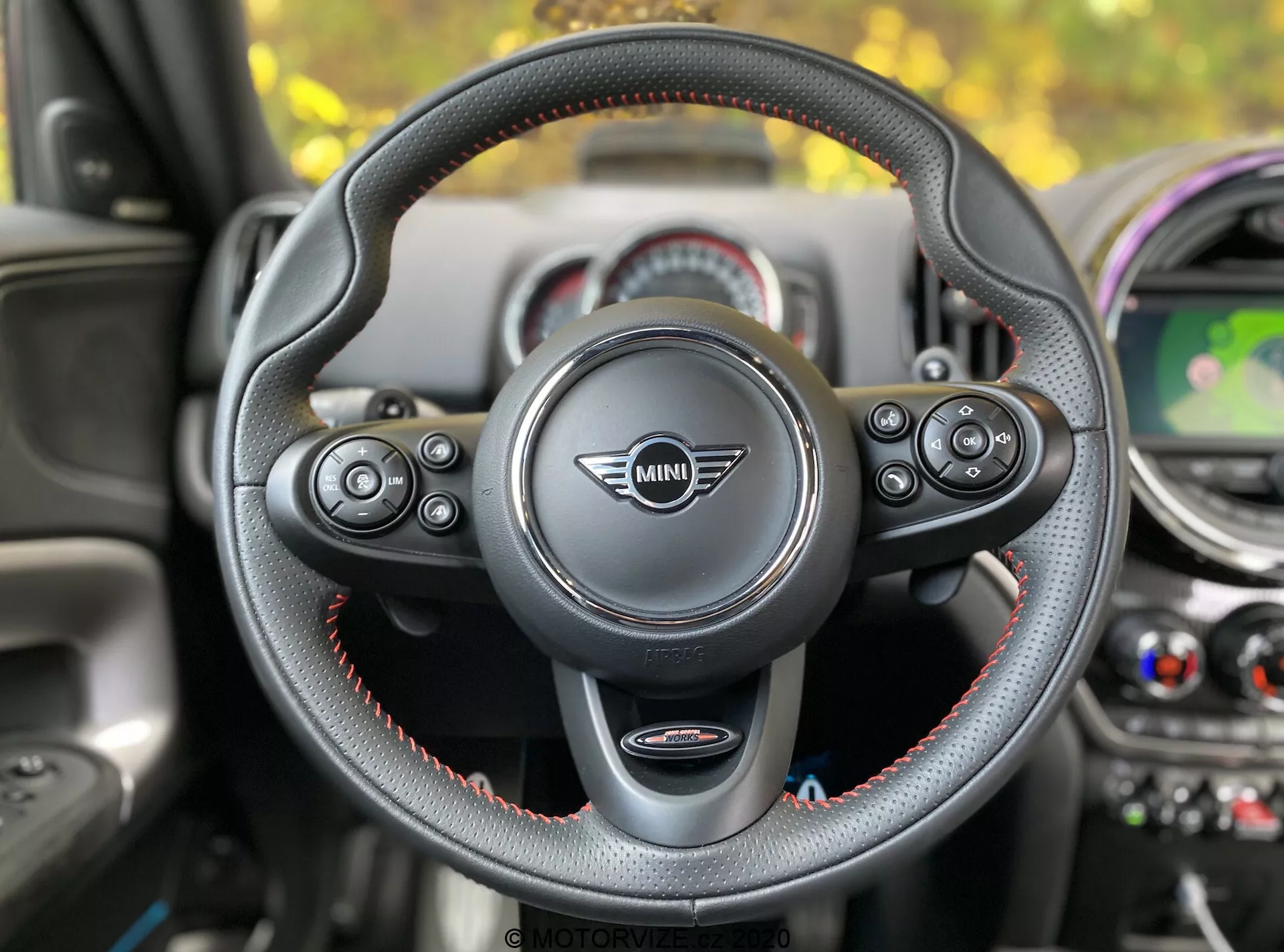 Pohľad do interiéru Mini Countryman John Cooper Works 2020 z miesta vodiča, zameraný na volant s logom Mini uprostred. Kožený trojramenný volant s červeným stehom má multifunkčné ovládanie pre audio, telefón, hlasové ovládanie a tempomat. Viditeľné sú páčky radenia, čo naznačuje automatickú prevodovku s manuálnym radením. V pozadí je prístrojová doska s kruhovými prvkami, centrálnym displejom zobrazujúcim mapu, vetracími otvormi a farebnými akcentmi.