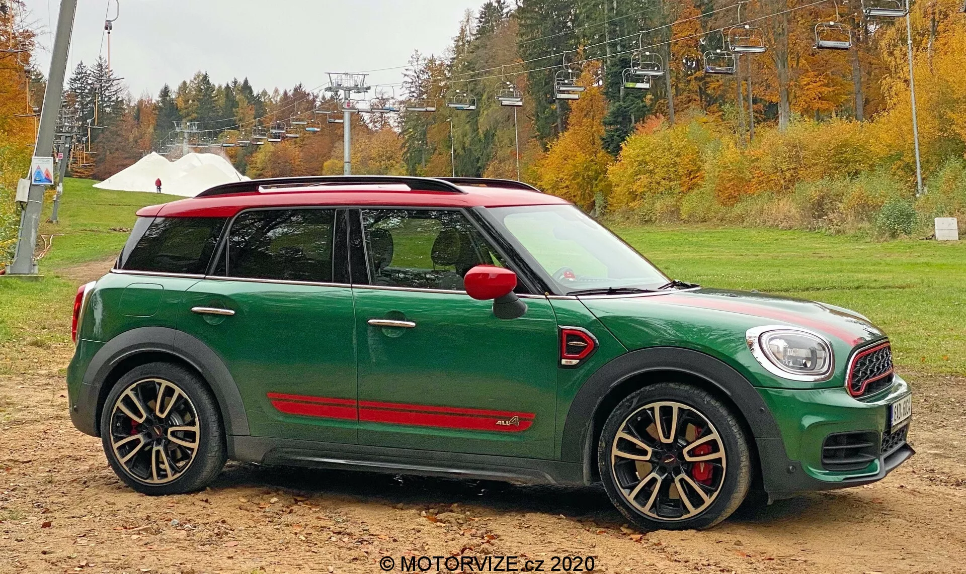 Bočný profil zeleného Mini Cooper Countryman 2020 John Cooper Works umiestneného na piesočnatej zemi s jesennými stromami a infraštruktúrou lyžiarskeho vleku v pozadí. Auto má bielu strechu, červené kryty bočných zrkadiel, agresívne čierno-strieborné kolesá s viacerými lúčmi s červenými brzdovými strmeňmi a športové červené bočné sukne.