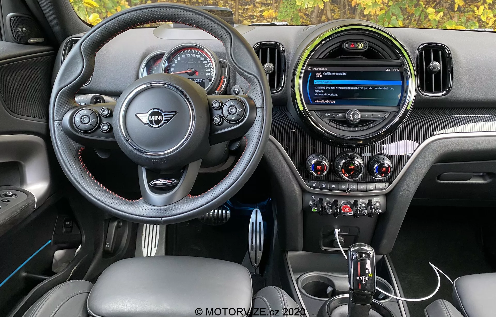 Interiérový pohľad na Mini Countryman John Cooper Works 2020, zvýrazňujúci kokpit vodiča so športovým volantom s plochým spodkom, multifunkčnými tlačidlami, kontrastným prešívaním a centrálnym emblémom výrobcu. Prístrojová doska zahŕňa moderný kruhový infotainment displej, tri kovové vzduchové výduchy a dobre integrované tlačidlá klimatizácie. Stredová konzola obsahuje automatickú radiacu páku s koženou manžetou a ovládacím gombíkom infotainmentu, doplnené okarbónovou textúrou, kožou a kovovými akcentmi pre prémiový a športovo ladený dizajn.