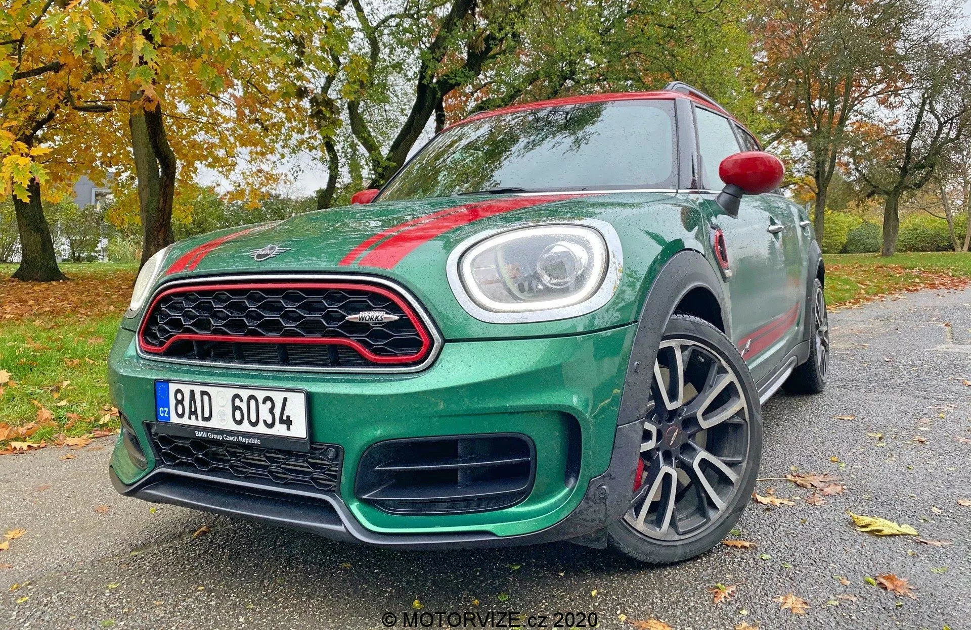 Obrázok zobrazuje hatchback Mini Countryman John Cooper Works 2020, zachytený z nízkeho predného trojštvrťového uhla. Viditeľné sú kľúčové dizajnové a technologické prvky, vrátane LED svetlometov, výraznej prednej mriežky, štýlového nárazníka, hliníkových kolies, čiastočne ukázaných spätných zrkadiel s integrovanými smerovkami, aerodynamických línií karosérie, odznakov a lesklého laku auta. Táto perspektíva zdôrazňuje agresívny postoj a dynamický dizajn vozidla.