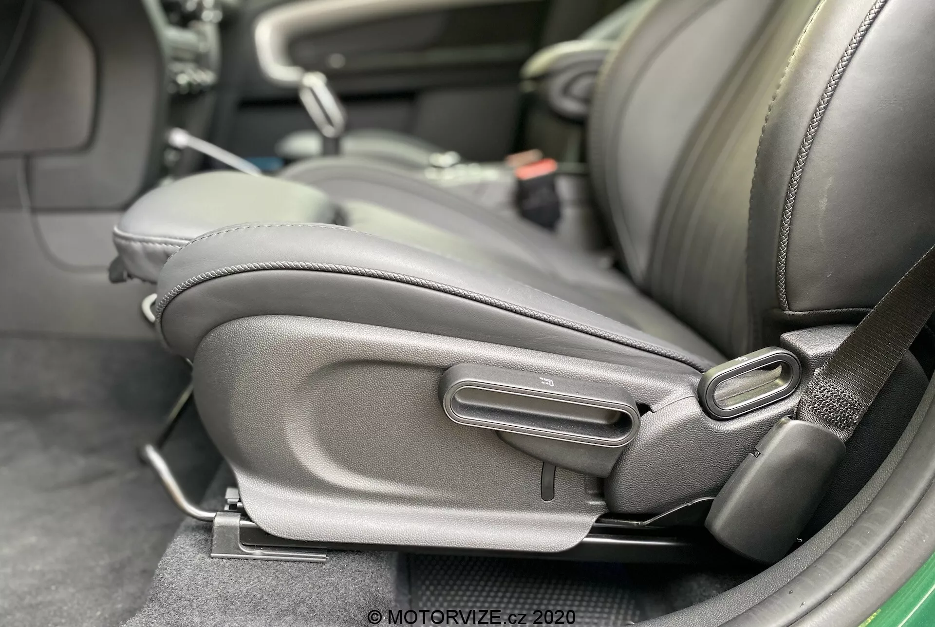 Detailný pohľad na interiér Mini Countryman John Cooper Works 2020, zameraný na spodnú bočnú časť vodičovho sedadla s detailnými mechanizmami nastavenia sedadla. Sedadlo je čalúnené tmavou kožou alebo kožešinou so zreteľným bočným opierkou, čo naznačuje dôraz na pohodlie a kvalitu. Viditeľné sú manuálne ovládacie prvky, vrátane rukoväti tyče a okrúhleho otočného voliča, čo naznačuje ergonomický dizajn a individuálne nastavenie. Prítomná je aj pracka bezpečnostného pásu, čo poukazuje na pozornosť venovanú bezpečnosti cestujúcich.