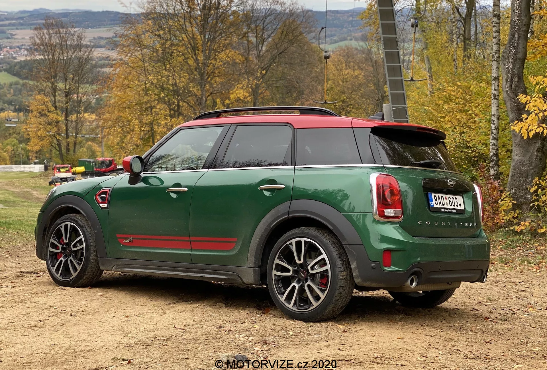 Trojštvrťový zadný pohľad na zelený Mini Countryman John Cooper Works 2020 s kontrastnou čiernou strechou a červenými bočnými zrkadlami. Viditeľné prvky zahŕňajú veľké viacramenné zliatinové kolesá so striebornými okrajmi, modelové označenie 'Cooper S', zaoblené zadné svetelné klastre s červenými a bielymi prvkami, zadný nárazník s parkovacími senzormi, dvojité výfukové rúrky, čierne strešné spojlery, chrómové kľučky dverí a červenú akcentnú líniu. Auto je zaparkované na nespevnenom povrchu s vidieckym pozadím trávy a stromov.