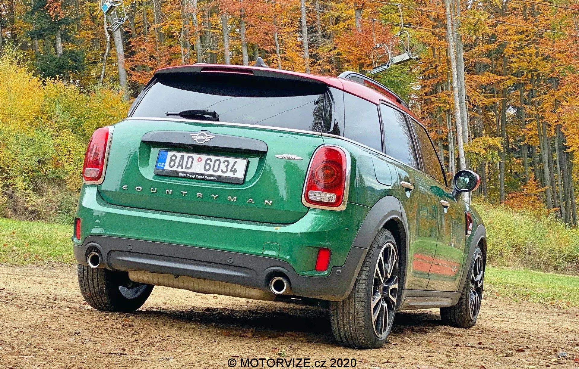 Obrázok zeleného Mini Countryman 2020 modelu John Cooper Works zaparkovaného vonku v jesennom prostredí, zhotovený zo zadného trojštvrťového uhla. Pravá zadná strana auta je otočená k fotoaparátu a ukazuje zadné svetlá, zadné okno, nápis 'COUNTRYMAN', dvojitý výfukový systém, päťlúčové kolesá, kontrastnú čiernu strechu a bočné zrkadlá. Obrázok tiež zahrňuje zadnú poznávaciu značku, zadný stierač a anténu pre konektivitu.