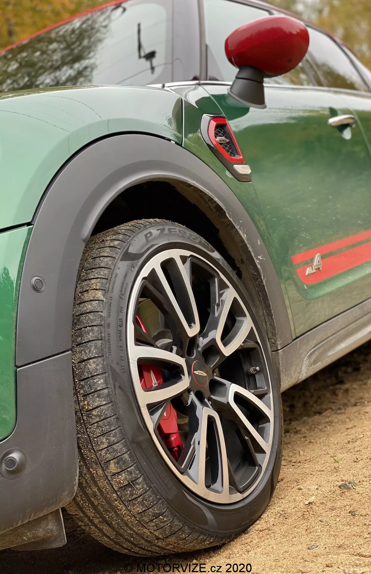 Detailný pohľad na zadné koleso a časť zadného blatníka Mini Countryman John Cooper Works 2020. Záber je urobený z bočného, mierne šikmého a nízkeho uhla, aby vyniklo viaclúčové zliatinové koleso v sivej alebo striebornej farbe. Červený brzdový strmeň naznačuje vysoko výkonný brzdový systém. Pneumatika má viditeľný dezén, čo naznačuje, že je vhodná pre rôzne cestné podmienky. Zelené auto má stylizovaný pruh pozdĺž spodnej časti dverí a čiastočne viditeľný emblém, čo pridáva športový vzhľad. Viditeľné je tiež zrkadlo s červeným krytom a zástavky, čo zvyšuje dynamický vzhľad vozidla. Rozmazané pozadie upriamuje pozornosť na tieto detaily, ideálne pre zdôraznenie prvkov výkonnosti automobilu.