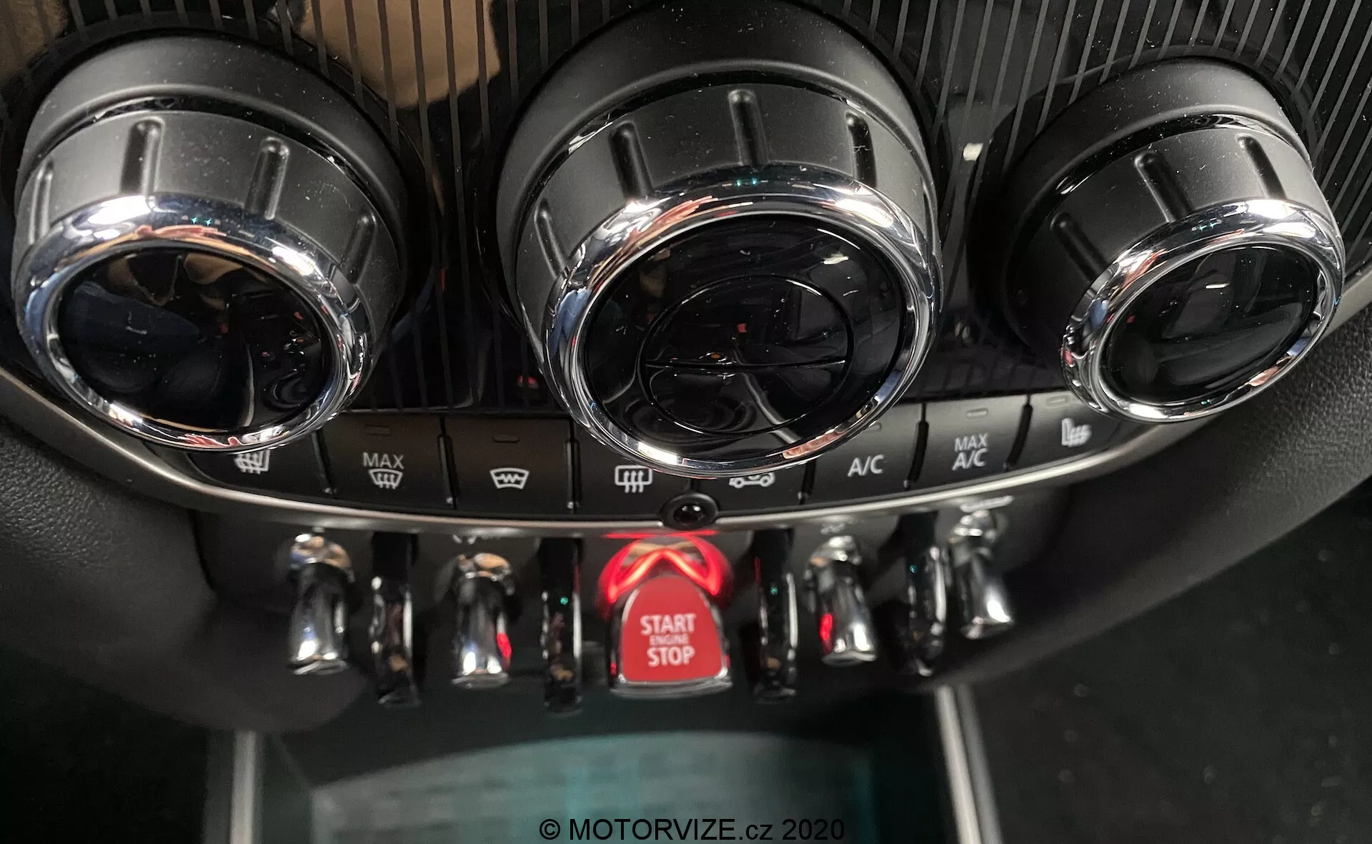 Tento obrázok zachytáva detailný pohľad na stredovú konzolu palubnej dosky v modeli Mini Countryman John Cooper Works z roku 2020, so zameraním na klimatizačný systém a tlačidlo štart-stop motora. Konzola zobrazuje dva kruhové vetracie otvory s kovovou povrchovou úpravou v hornej časti, podrobný panel klimatizácie s tromi kovovými otočnými ovládačmi pre nastavenie teploty, rýchlosti ventilátora a smeru prúdenia vzduchu a niekoľkými tlačidlami pre funkcie ako odmrazovanie a recirkulácia vzduchu. Pod ovládacími prvkami sa nachádza výrazné červené tlačidlo na spúšťanie a vypínanie motora. Textúry panelu zahŕňajú športový dizajn pripomínajúci uhlíkové vlákno okolo vetracích otvorov a štandardný čierny plast pre spodný panel. Uhol fotografie, z prednej strany a mierne zhora, zvýrazňuje vysoko kvalitný dizajn a pokročilé funkcie.