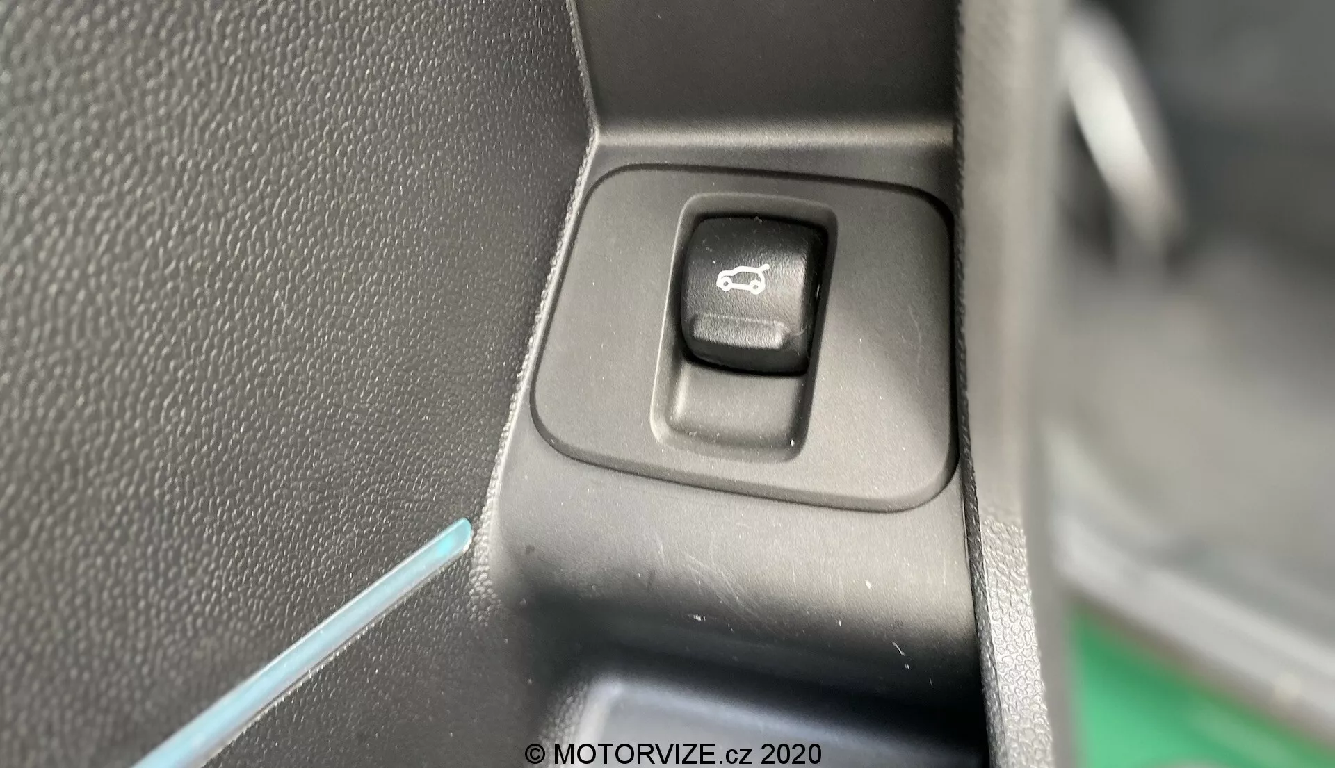 Detailný pohľad na ovládací spínač na ovládanie bočných zrkadiel v interiéri Mini Countryman 2020 John Cooper Works. Obdĺžnikový spínač so zaoblenými rohmi, umiestnený na plochom paneli, je zvýraznený symbolom zrkadla. Jedna strana spínača je v tieni, čo naznačuje svetelný zdroj z opačnej strany. Pozadie tvorí čierny textúrovaný plast a tenká modrá čiara ambientného osvetlenia, čo prispieva k estetike interiéru vozidla.