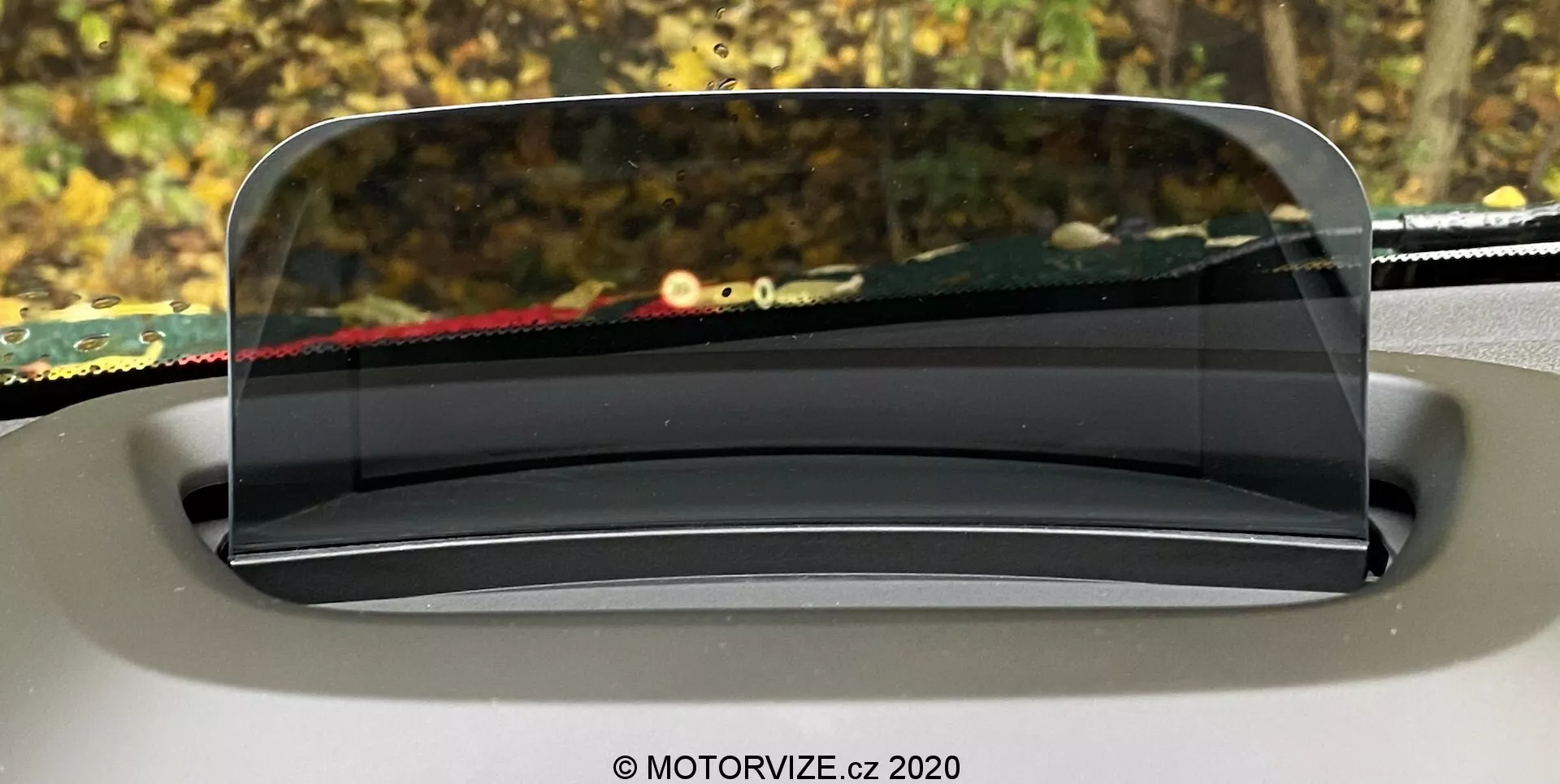 Obrázok zobrazujúci head-up displej (HUD) vozidla Mini Countryman 2020, premietaný na priehľadnú obrazovku nad prístrojovou doskou z perspektívy vodiča. Priehľadný, sklený panel HUD je plynulo integrovaný s interiérom, zaoblený a mierne naklonený pre optimálnu viditeľnosť. Pozadie za čelným sklom je rozmazané, čo zdôrazňuje zameranie na HUD a zobrazovanie dôležitých informácií pre vodiča.