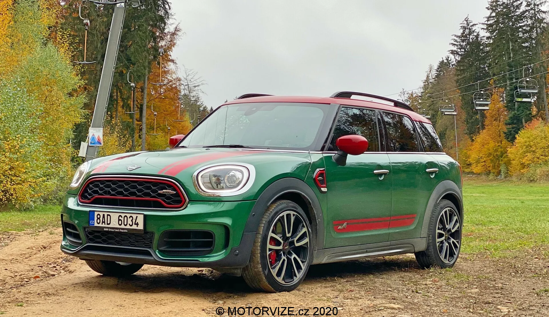Obrázok zeleného Mini Countryman John Cooper Works 2020 v terénnej štrkovej oblasti s lesnou krajinou. Kompaktný crossover zobrazený v trojštvrťovom prednom uhle s kontrastnou červenou strechou a bočnými zrkadlami, veľkou prednou mriežkou s chrómovým lemom, LED denným svietením obklopujúcim hlavné svetlomety a hmlovkami integrovanými do spodného nárazníka. Výrazné športové prvky zahŕňajú kapotu s prívodom vzduchu, čierne oblúky kolies, bočné prahy, zadný spoiler, veľké viacramenné zliatinové disky s nízkoprofilovými pneumatikami a červené brzdové strmene. Viditeľné sú aj panoramatická strecha a aerodynamické strešné lišty.