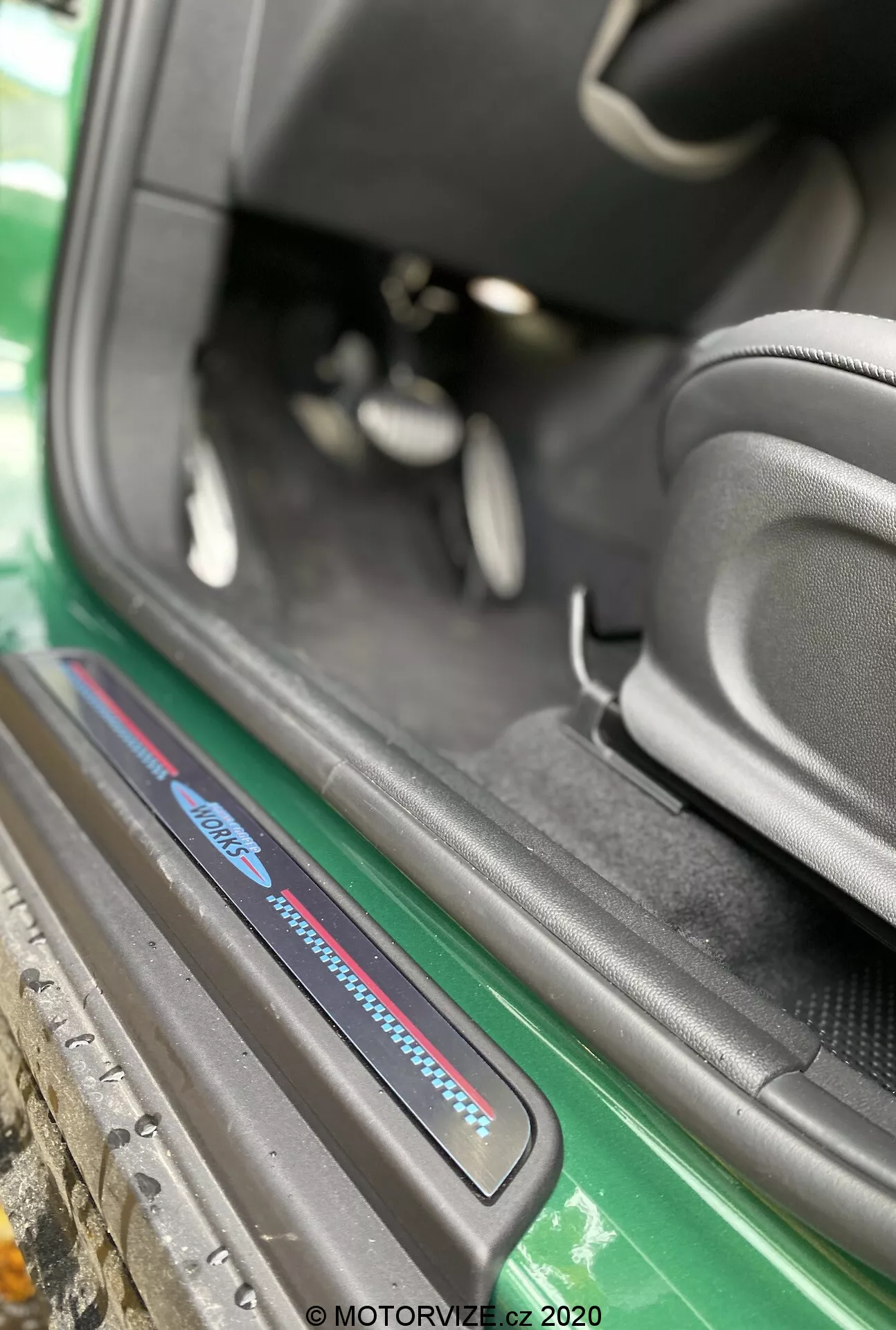 Detailný pohľad na oblasť prahu dverí Mini Countryman John Cooper Works 2020 z úhlopriečneho uhla zameraného na prah na strane spolujazdca. Obrázok, zachytený zvonka vozidla, zdôrazňuje spodnú časť interiéru vozidla vrátane prahovej dosky s modro-červeným logom, čierne koberce, športové bočné sedadlo, čiastočný pohľad na pedále a dobre udržiavanú oblasť prahu. Viditeľné vonkajšie prvky zahŕňajú zelenú bočnú sukňu a karosériu, ktoré zvyšujú estetický dojem. Vodotlač 'MOTORVIZE.cz 2020' naznačuje, že fotografia bola urobená v roku 2020 pre automobilovú recenziu.