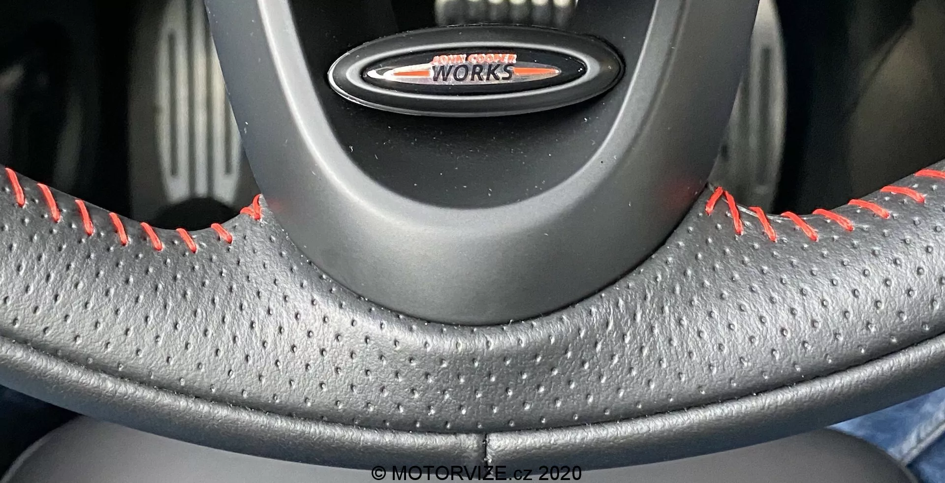 Detailný pohľad na volant z perspektívy vodiča, s emblémom 'John Cooper Works', koženým poťahom s textúrou pre lepší úchop a červeným kontrastným šitím typickým pre výkonné varianty MINI Cooper