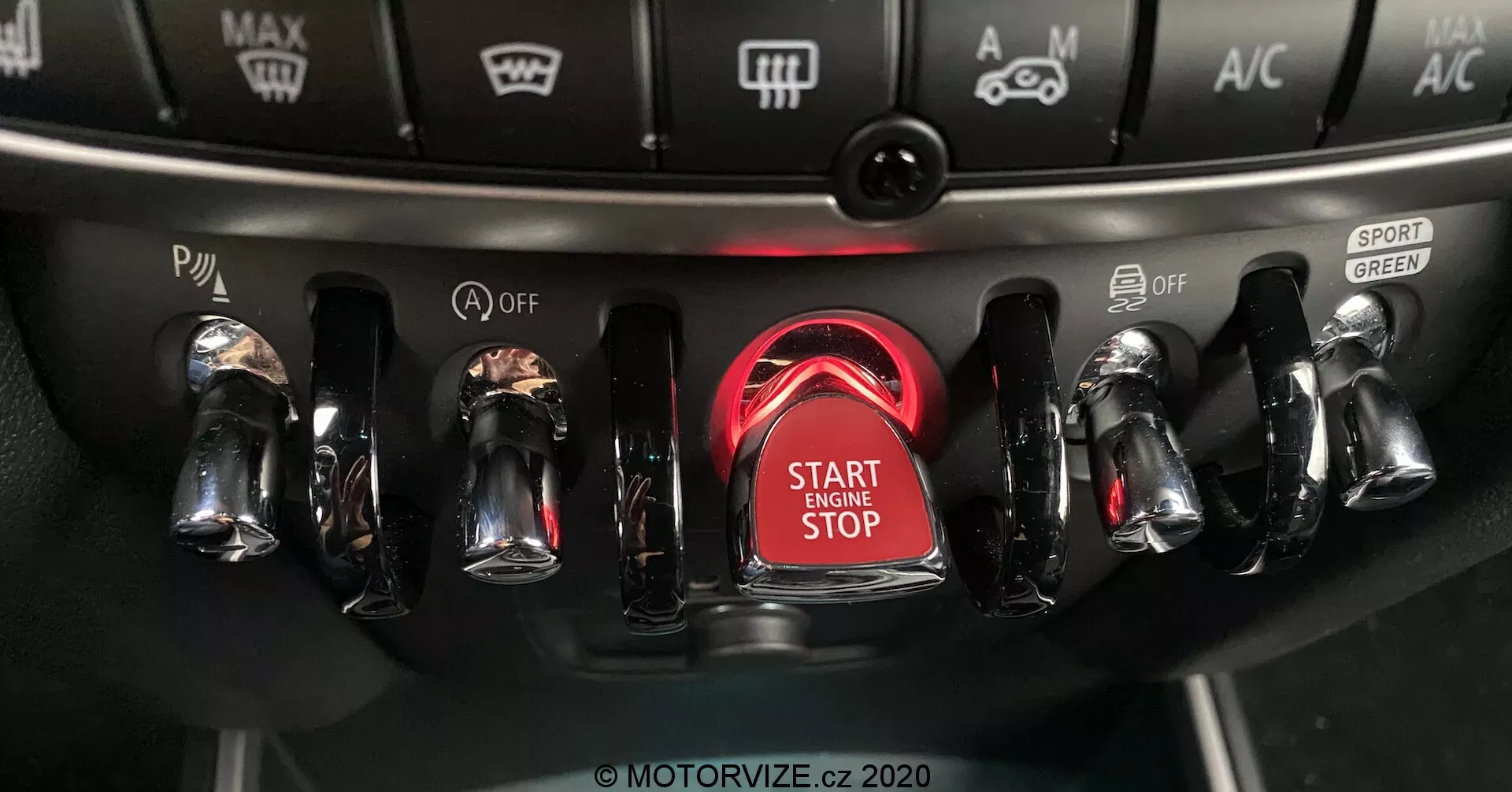 Detailný pohľad na stredovú konzolu Mini Countryman John Cooper Works 2020 so zameraním na oblasť zapaľovania s výrazne zvýrazneným tlačidlom štart/stop motora obklopeným žiariacim červeným kruhom, obsahujúcim kovové prepínače s chrómovaným povrchom na oboch stranách pre rôzne ovládacie prvky vozidla vrátane jazdných režimov a nastavení klimatizácie.