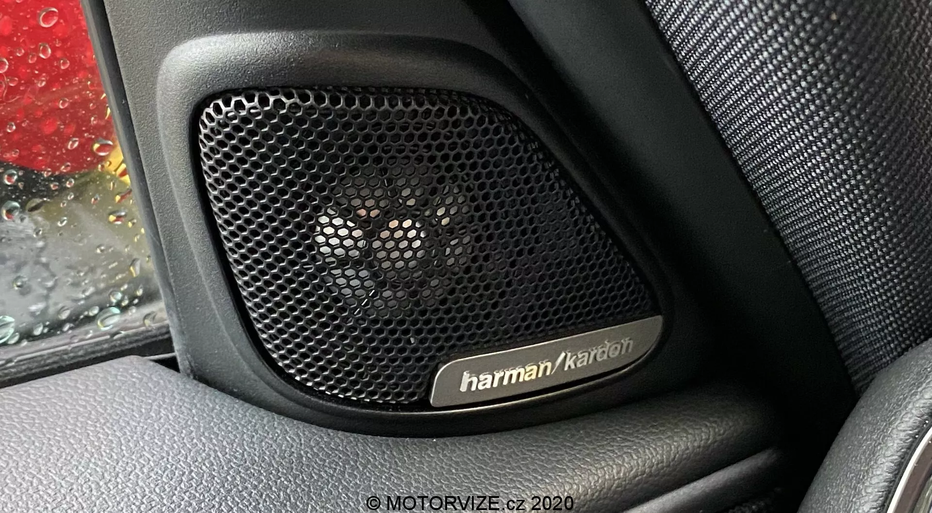 Detailný pohľad na audiosystém Harman Kardon vnútri dverí vozidla Mini Countryman 2020 John Cooper Works. Fotografia sa zameriava na perforovanú mriežku reproduktora, navrhnutú na akustickú priehľadnosť a ochranu, zasadenú do interiéru s textúrou podobnou koži. Na okne auta sú viditeľné kvapky dažďa, čo dodáva kontext snímke. Prémiový audiosystém poukazuje na kľúčové technologické prvky, ukazujúc luxus a odolnosť dizajnu vozidla.