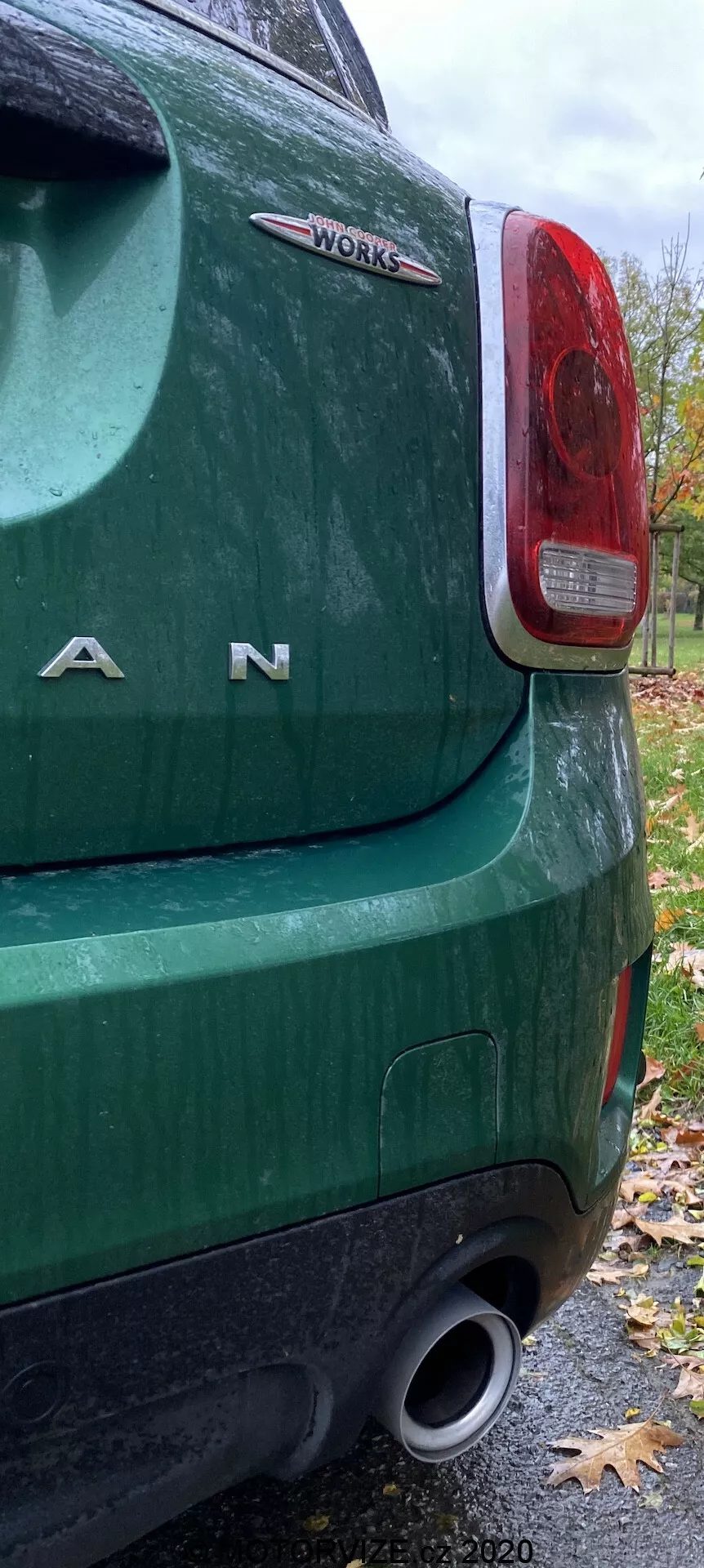 Detailný pohľad na zadný ľavý roh zeleného 2020 Mini Countryman John Cooper Works so zameraním na zadné svetlo, chrómové písmená 'MINI', odznak 'WORKS', zadný nárazník a výfuk, čo naznačuje vysokovýkonné prvky. Obrázok je urobený za zamračeného jesenného dňa s mokrým povrchom a opadanými listami.