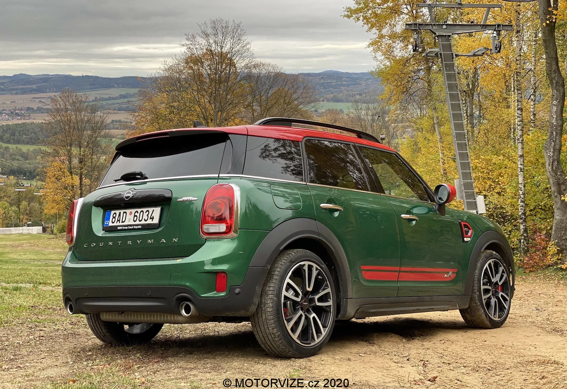 Trojštvrťový zadný pohľad na zelený Mini Cooper Countryman John Cooper Works 2020 zaparkovaný vonku, zvýrazňujúci dizajn auta z bočného zadného pohľadu. Významné detaily zahŕňajú tmavozelenú karosériu s červenou strechou, odznak 'Countryman', logo Mini na zadnom dverách, charakteristické zadné svetlá, dvojitý výfukový systém, zliatinové kolesá, panoramatickú strechu, zadný stierač, tónované zadné okná, parkovacie senzory a bočné zrkadlo s integrovaným smerovkou. Vozidlo je na nespevnenom povrchu s jesenným lístím a štruktúrou lyžiarskeho vleku v pozadí, čo naznačuje aktívny, vonkajší životný štýl.