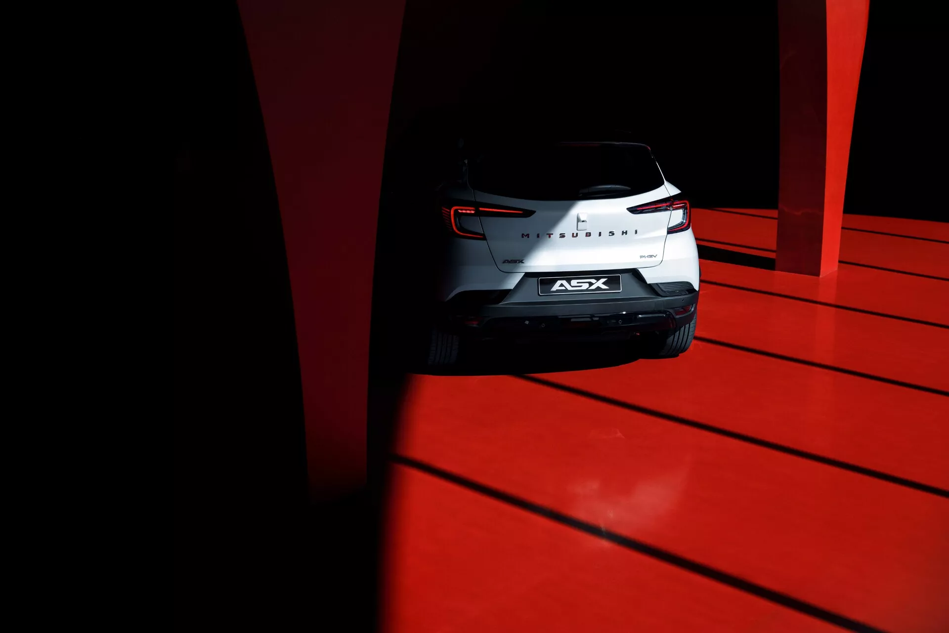 Nízky uhol záberu na zadnú časť kompaktného SUV Mitsubishi ASX 2023 s vypracovanými zadnými svetlami, chrómovým znakom a zadným sklom, zachytený za vysokého kontrastu denného svetla na žiarivo červenom povrchu.