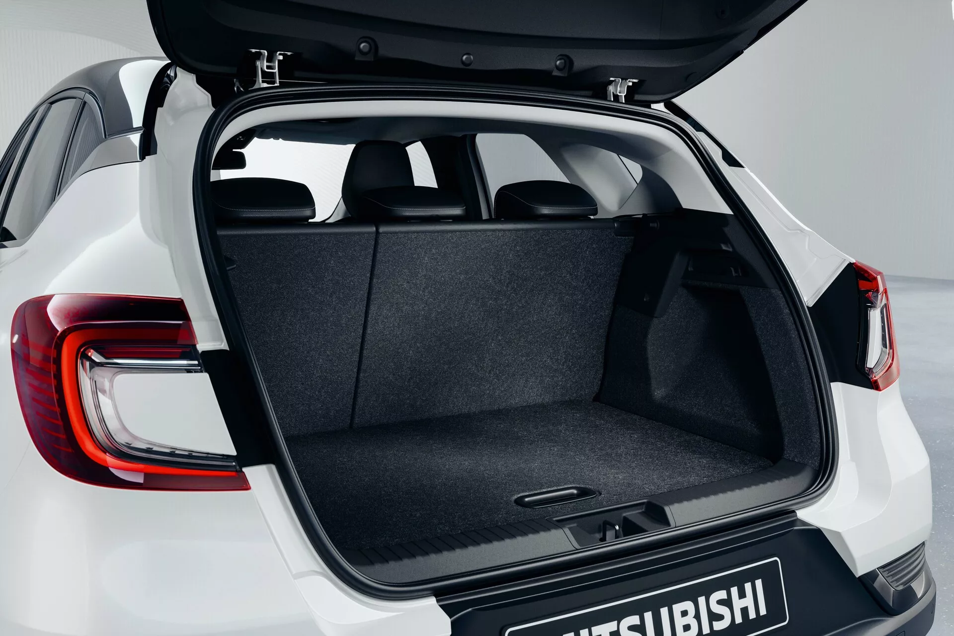 Zadný priestor modelu Mitsubishi ASX 2023 (2. generácia) s otvoreným kufrom z mierne zvýšeného uhla, zvýrazňujúci kobercový priestor, mechanizmus pántov, rovné nakladacie dno a čiastočne viditeľné moderné LED zadné svetlá s logom Mitsubishi na dverách kufra.