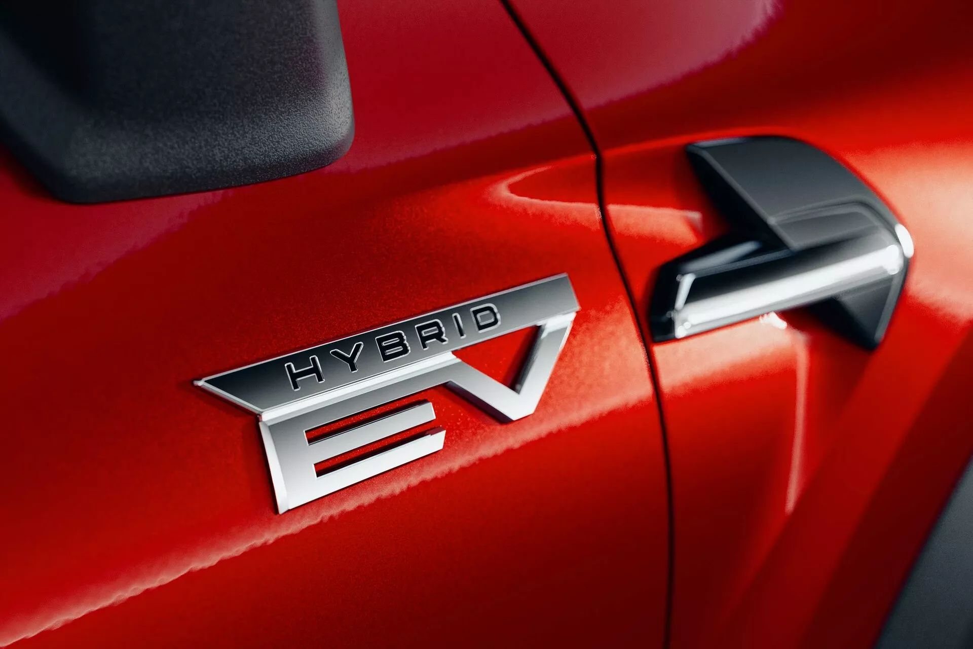 Detailný záber na Mitsubishi ASX 2023 (2. generácia) v červenej farbe, ukazujúci výrazný odznak 'HYBRID EV', ktorý naznačuje technológiu hybridného elektrického vozidla. Odznak 'HYBRID EV' je umiestnený v strede, zdôrazňujúc kovovú úpravu a presné písmo. 'EV' má výrazný dizajn so šípkou prechádzajúcou cez 'V'. Bočné zrkadlo je čiastočne viditeľné v pravom hornom rohu, čo naznačuje prítomnosť ďalších technologických prvkov. Lesklá povrchová úprava laku a tesný šev medzi prednými dvermi pasažiera a predným blatníkom naznačujú vysokú kvalitu spracovania.