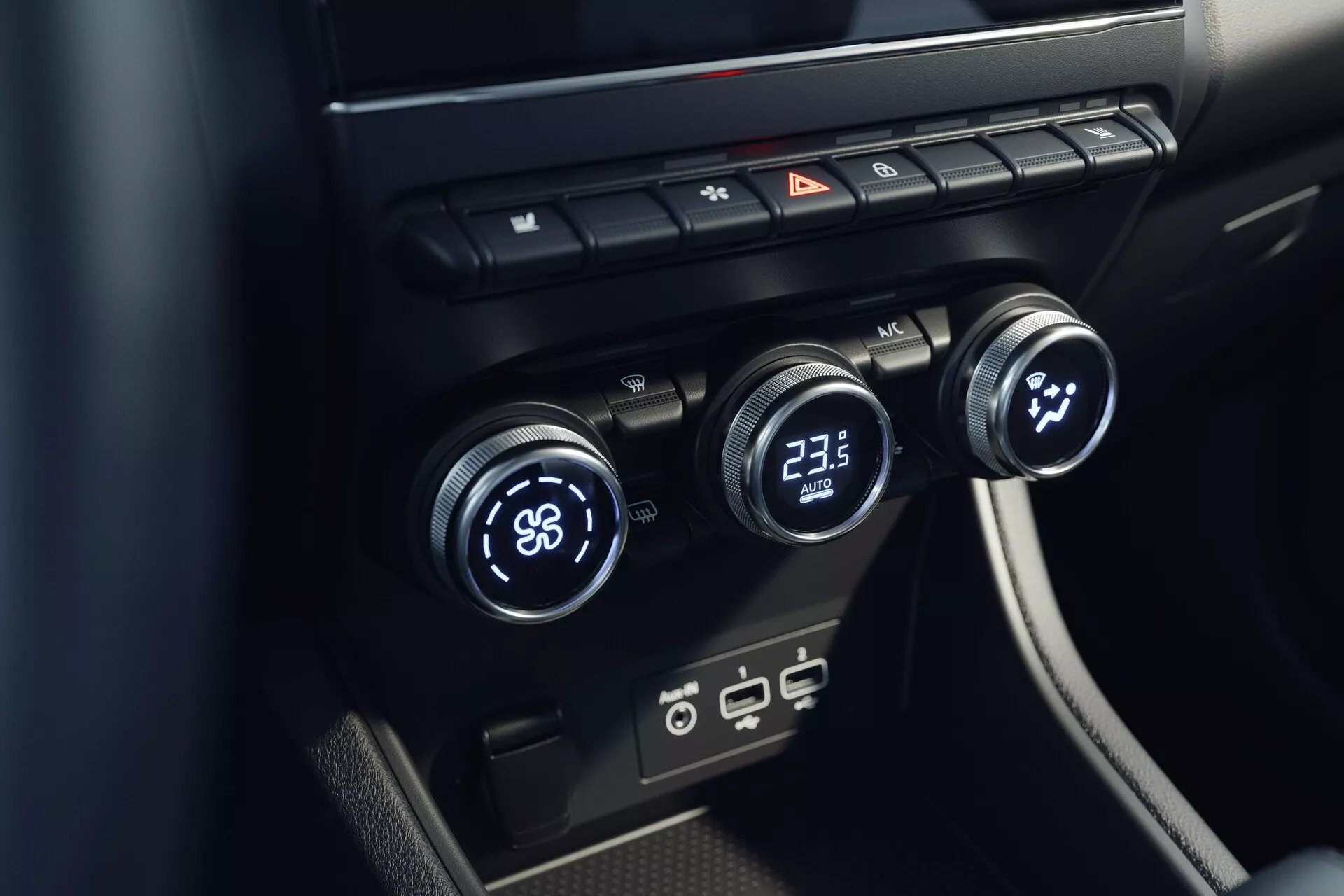 Detailný pohľad na systém klimatizácie interiéru Mitsubishi ASX 2023 (2. generácia). Obrázok z vyššieho uhla sa zameriava na centrálnu konzolu s tromi ovládačmi teploty - ľavý nastavený na 'LO' a pravý na '23,5'. Stredný ovládač pravdepodobne riadi rýchlosť alebo smer ventilátora. Hore sú tlačidlá na odhmlievanie, výstražné svetlá a cirkuláciu vzduchu. Nižšie sú moderné USB porty. Dizajn je minimalistický s elegantnými čiernymi povrchmi.