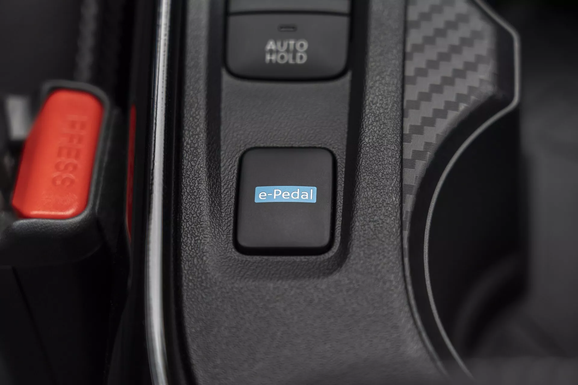 Detailný záber interiéru Nissan Juke (F16) 2019 so zameraním na spínač systému e-Pedal pre ovládanie elektrického alebo hybridného pohonu, s viditeľným tlačidlom AUTO HOLD a červenou sponou bezpečnostného pásu, čo naznačuje pokročilú automobilovú technológiu a prémiové vnútorné prvky
