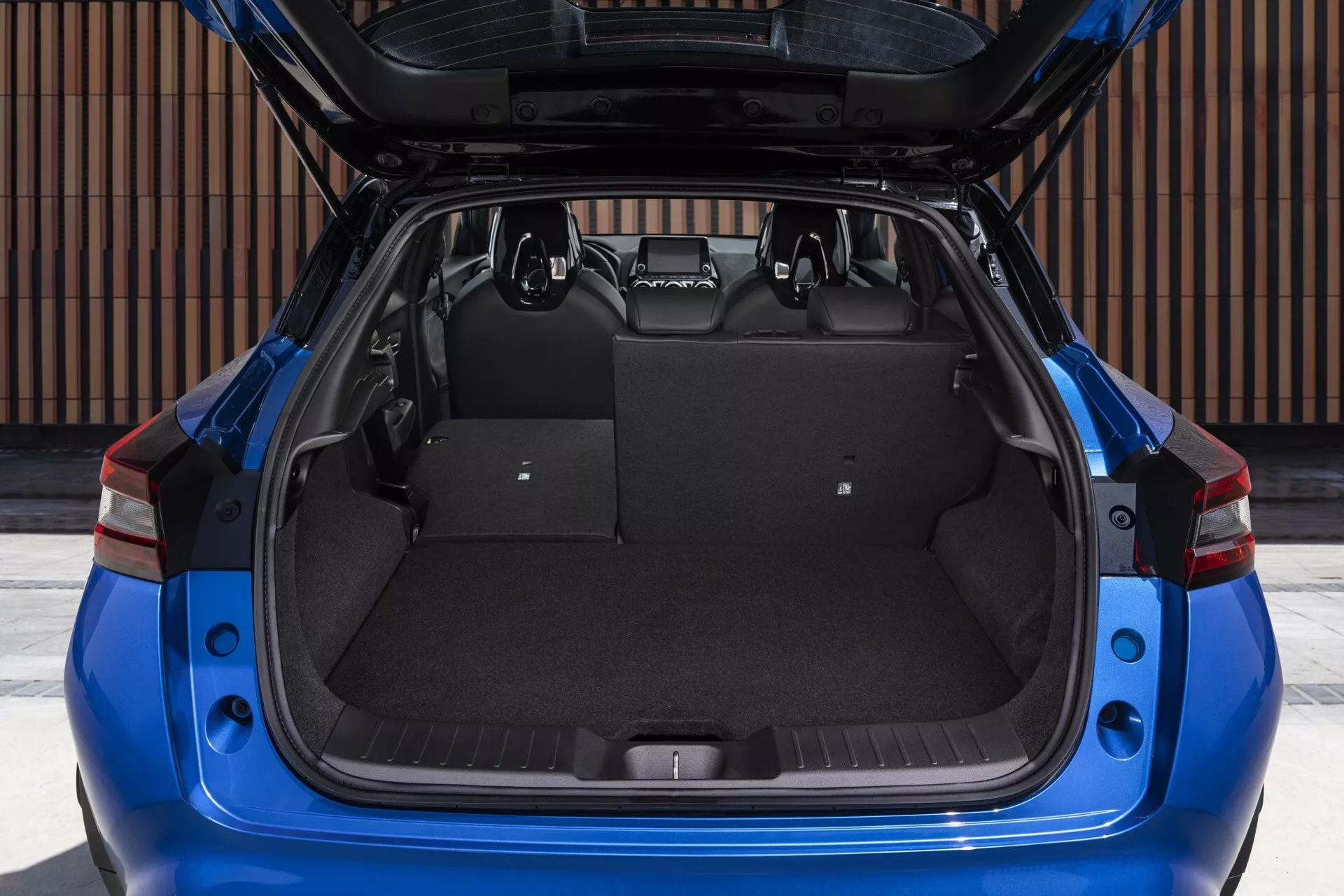 Nissan Juke (F16) z roku 2019 ukazuje priestranný a prázdny kufor s otvoreným vekom, detailný pohľad do nákladového priestoru s kobercom podobným čalúnením, orámovaný charakteristickými červenými zadnými svetlami a modernými prvkami vonkajšieho dizajnu, z priamej zadnej perspektívy.