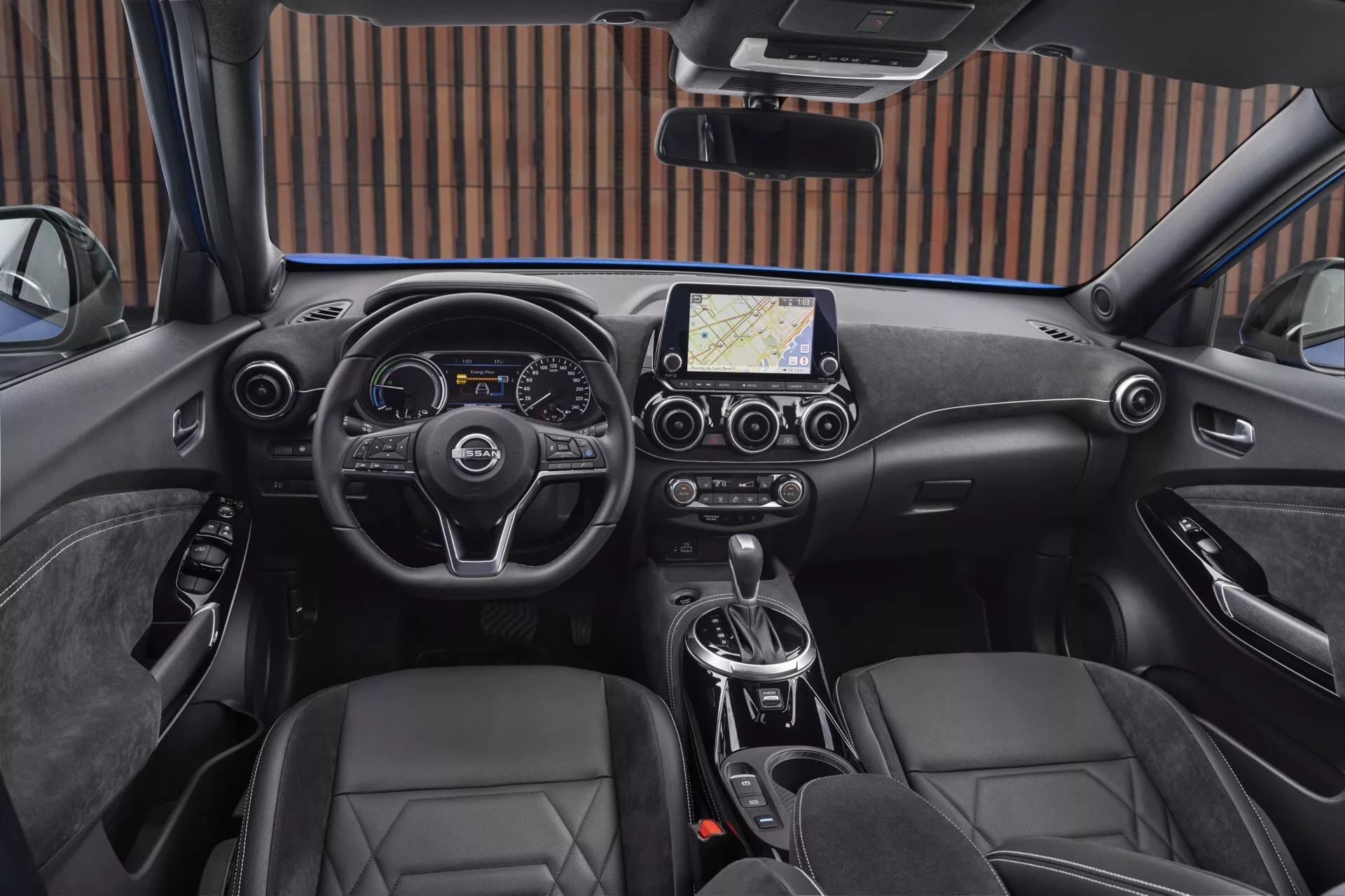 Interiér Nissan Juke F16 z roku 2019 z pohľadu vodiča, zahŕňajúci moderný volant s tromi ramenami s ovládacími tlačidlami, kombinovaný digitálny a analógový prístrojový panel, centrálny dotykový informačný systém s navigáciou, klimatizáciu s dvojzónovou reguláciou, automatickú prevodovku, sedadlá typu bucket potiahnuté kožou a elektronickú parkovaciu brzdu.