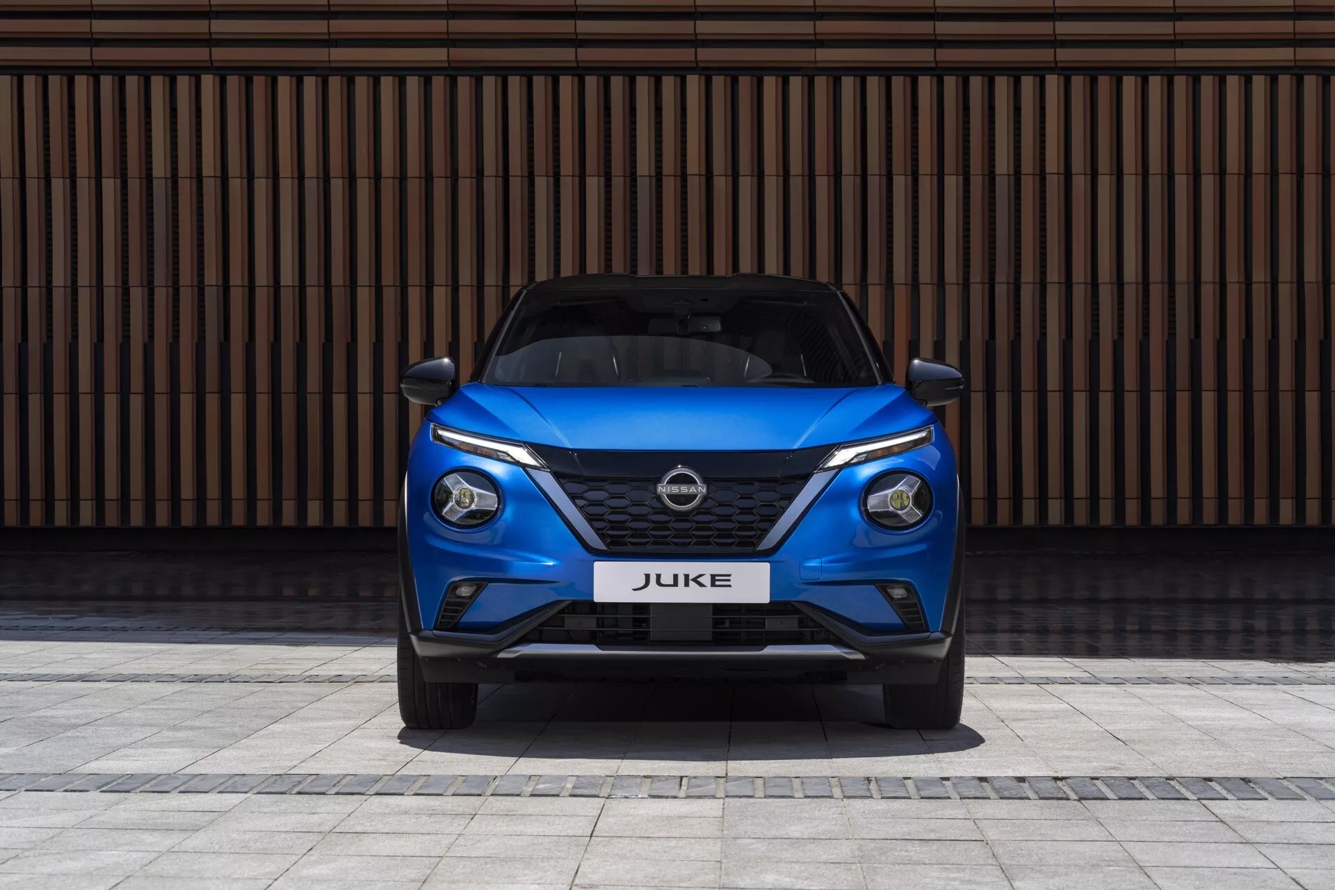 Predný pohľad na modrý Nissan Juke F16 z roku 2019, predvádzajúci elegantné LED svetlomety, mriežku V-motion a športový nárazník na pozadí so stenou s horizontálnymi čiarami