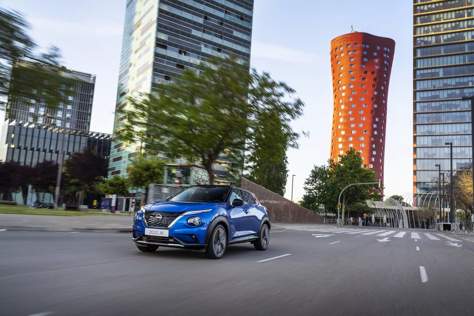 Nissan Juke F16 2019 v žiarivo modrej farbe, v pohybe na mestskom ceste s dynamickým pohľadom z prednej trojštvrte z pohľadu vodiča, predvádzajúcim zliatinové kolesá, tvarované karosérske línie a zapnuté svetlomety na pozadí moderného mesta s efektom rozmazania pohybu.
