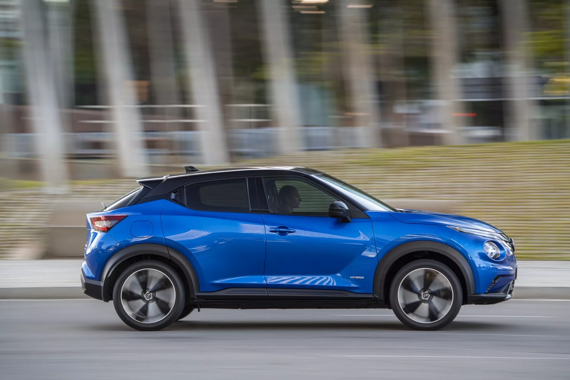 Modrý Nissan Juke (F16) z roku 2019, ukazujúci moderný dizajn a technologicky pokročilé funkcie ako aerodynamické línie karosérie, zliatinové kolesá a potenciálne bezkľúčové vstupné technológie, dynamicky sa pohybujúci s rozostrením pohybu počas zamračeného dňa