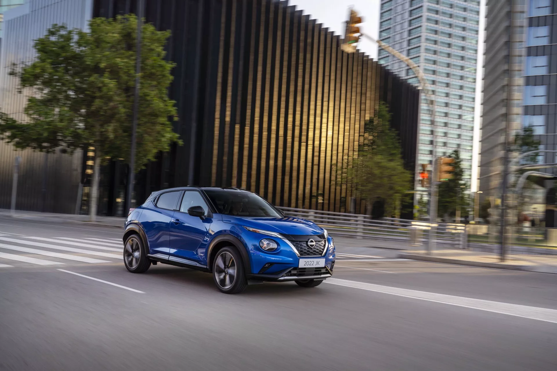 Moderné kompaktné SUV Nissan Juke (F16) z roku 2019 v pohybe, zachytené z bočnej perspektívy v jasne modrej farbe, ktoré zvýrazňuje dizajn vozidla, dynamickú prítomnosť a aerodynamické vlastnosti na mestskom pozadí.