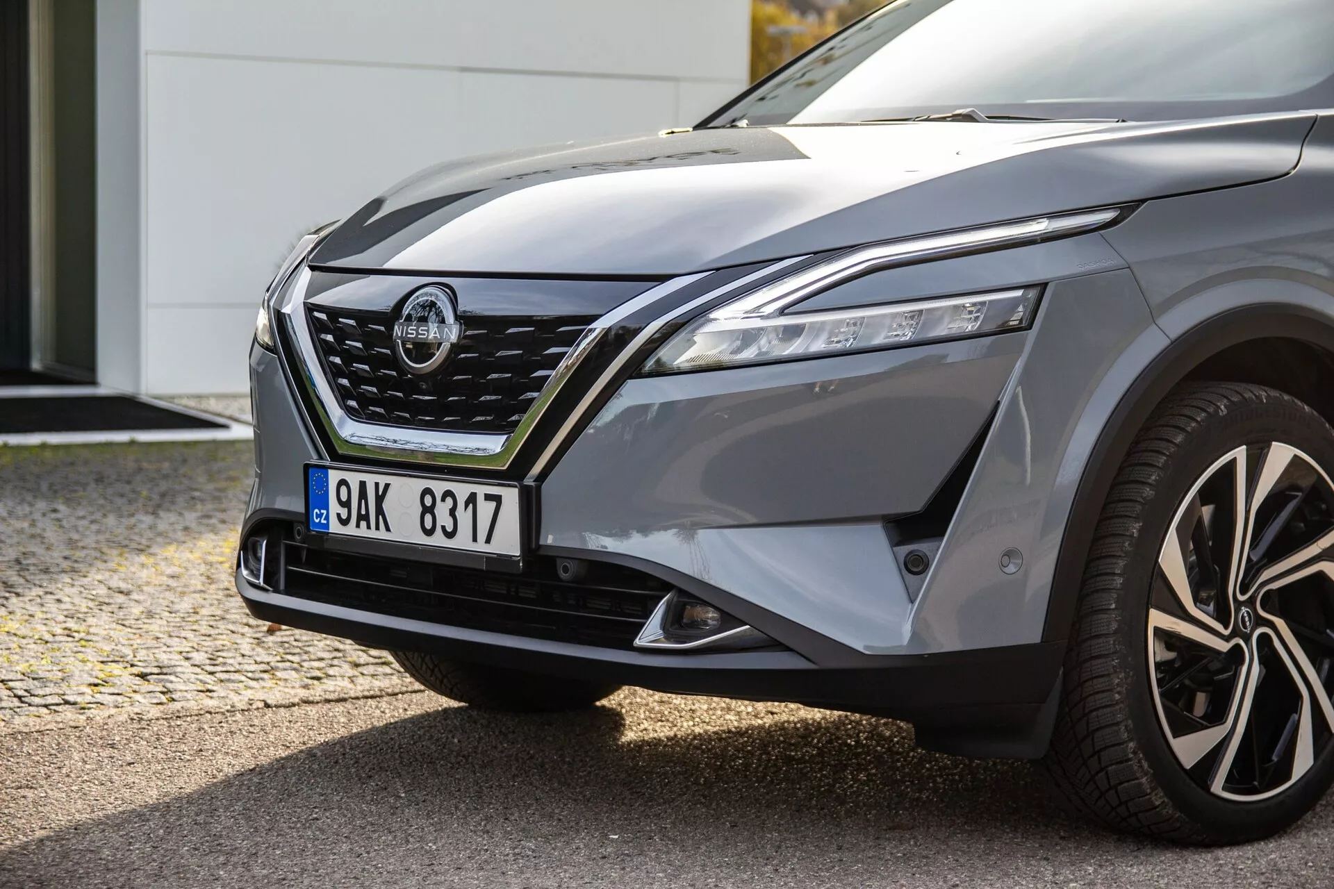 Detailný pohľad na prednú ľavú stranu moderného auta, ukazujúci elegantné LED svetlá, výraznú masku s chrómovými akcentmi, tvarovanú kapotu, vypuklý blatník, viaclúčové zliatinové kolesá a vodičove bočné spätné zrkadlo s integrovanými smerovkami, odrážajúc dynamický dizajn a potenciálne schopnosti ovládania auta.