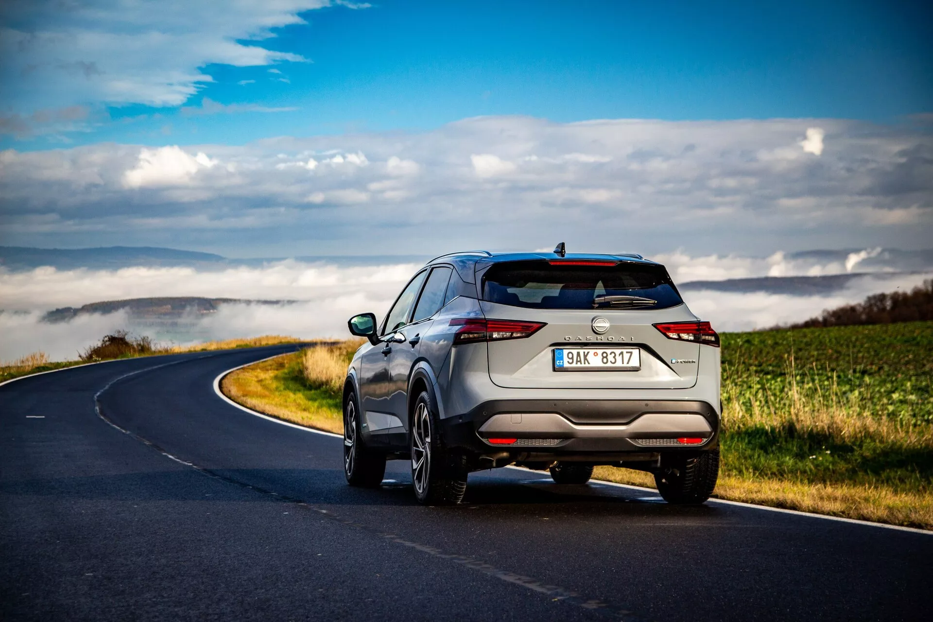 SUV Nissan Qashqai 2021 zachytený zo zadného trojštvrťového uhla na kľukatej vidieckej ceste pod rozptýlenými oblakmi. Dominujú moderný a elegantný dizajn s horizontálnymi úzkymi zadnými svetlami, diskrétnymi výfukovými rúrkami, viacramennými zliatinovými kolesami, strešnými lištami a pokročilými parkovacími senzormi. Svetlá metalická farba podčiarkuje jeho prémiový vzhľad.