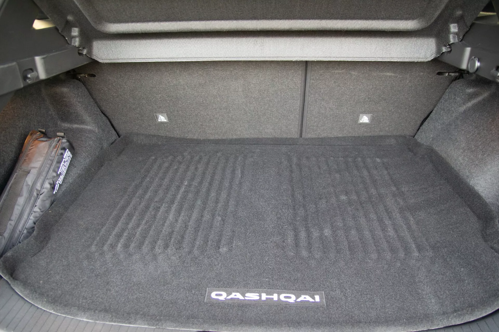 Obrázok zobrazujúci otvorený batožinový priestor Nissan Qashqai 2021 zo zadného uhlu. Prázdny kufor odhaľuje koberec, bočné steny a vzpriamené zadné sedadlá. Mierne zvýšená plocha na prednej strane kufru naznačuje mechanické komponenty alebo oporu pre zadné sedadlá. Vzadu je viditeľný menší úložný oddiel. Na ľavej strane je núdzové vybavenie, pravdepodobne zdvihák na pneumatiky alebo lekárnička. Obrázok zdôrazňuje priestrannosť a funkčný dizajn batožinového priestoru vozidla.