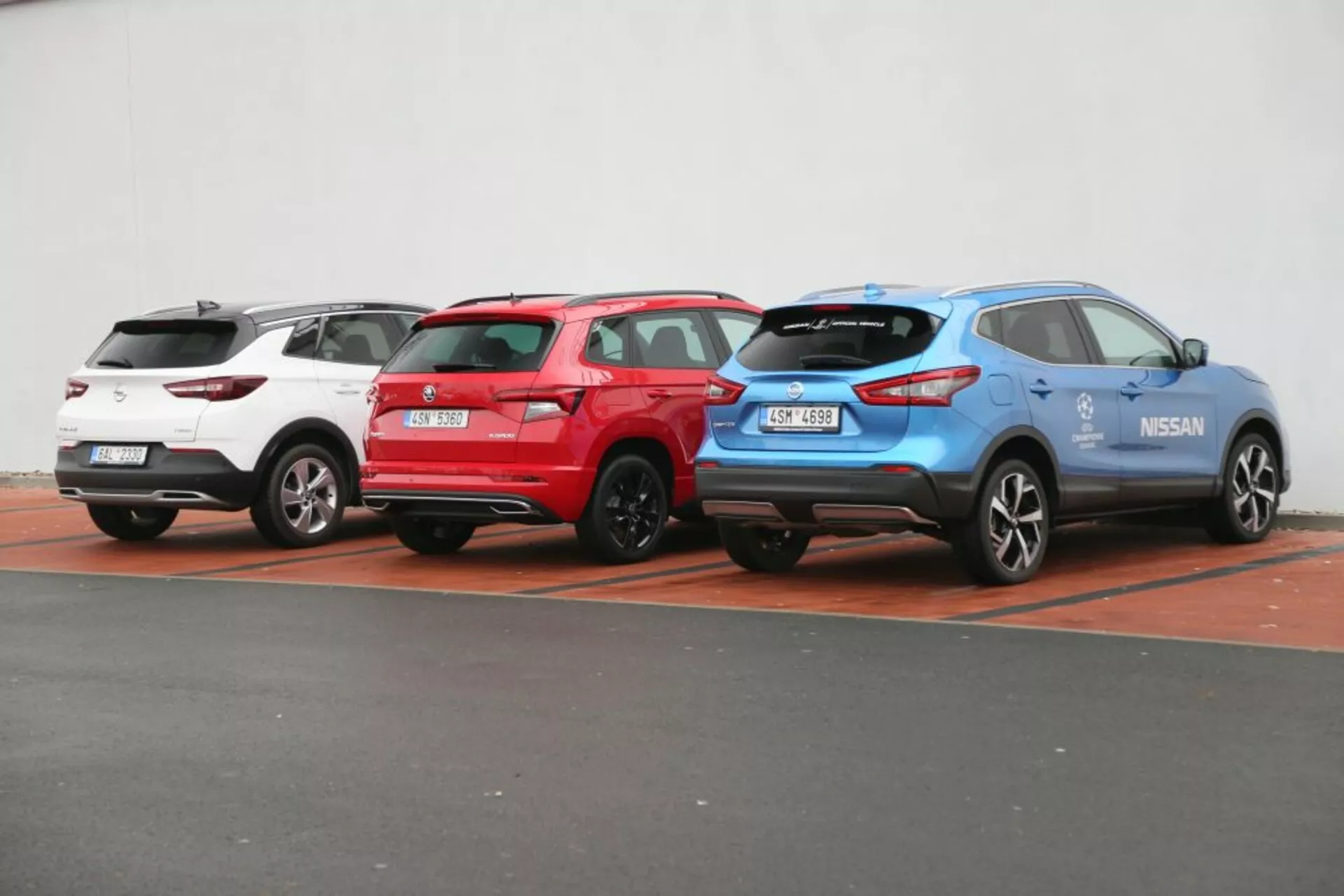 Tri SUV-ky Nissan Qashqai 2014 v bielej, červenej a modrej farbe zaparkované vedľa seba, zobrazujúce zadné a bočné profily s viditeľnými zadnými svetlami, stieračmi zadného skla a športovými zliatinovými kolesami na neutrálnom pozadí.