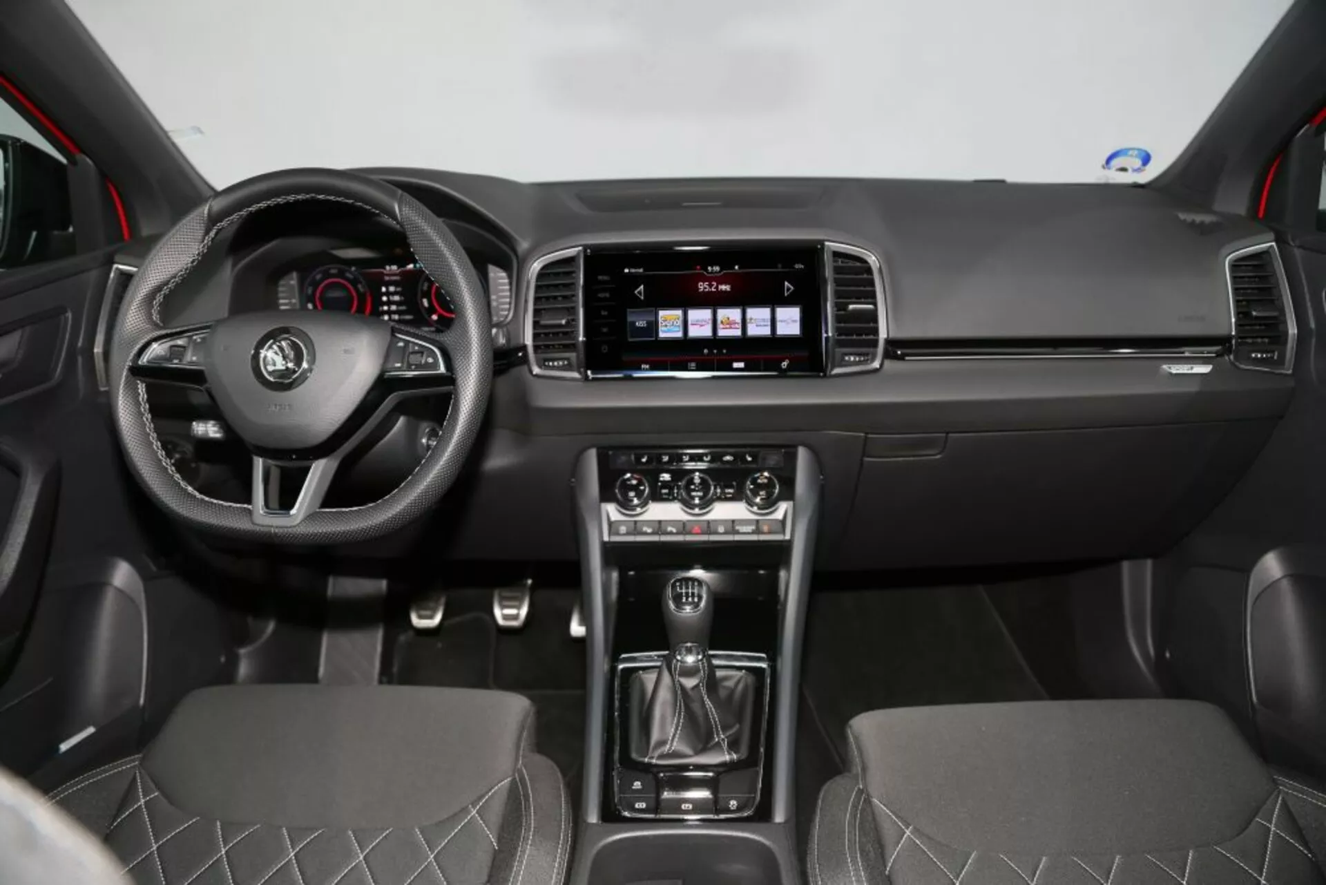 Čelný pohľad na interiér Nissan Qashqai 2014 ukazuje palubnú dosku, volant s ovládacími prvkami, systém infotainment, manuálnu radicu páku, klimatizáciu a sedadlá potiahnuté látkou s diamantovým prešívaním.