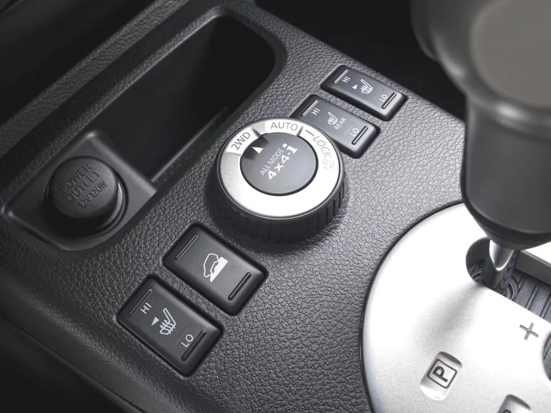 Detailný pohľad na stredovú konzolu Nissan X-Trail 2010 s ovládacím kolieskom 4WD, tlačidlami na vyhrievanie sedadiel, tlačidlom pre elektrickú zásuvku a radiacou pákou s možnosťou manuálneho radenia