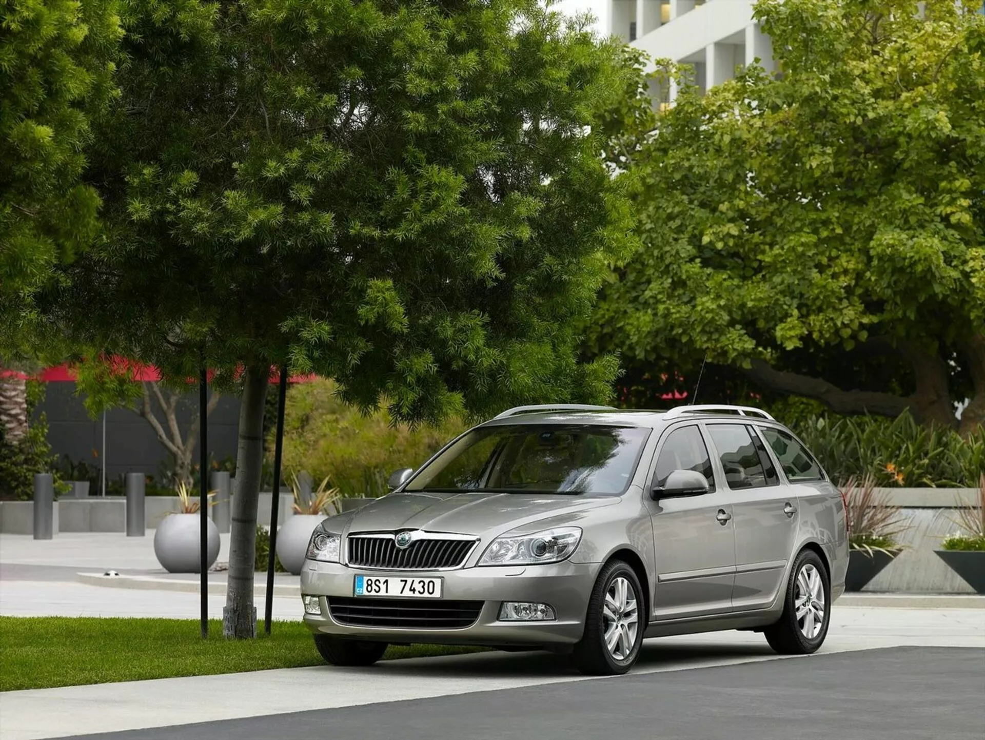 Station wagon Opel Astra z roku 2011 zaparkovaný v mestskom prostredí so zeleňou a modernými budovami na pozadí, predvádzajúci prednú mriežku vozidla, svetlomety a elegantný bočný profil vrátane zliatinových kolies a strešných reliéfov