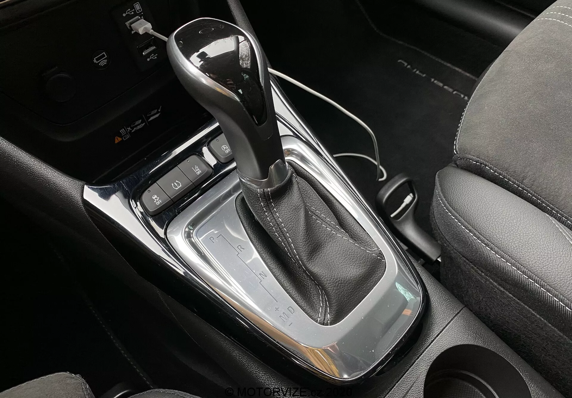 Detailný pohľad na stredovú konzolu v modernom modeli Opel Crossland 2021, zvýrazňujúci automatickú radiacu páku zabalenú v čiernej koži s chrómovým lemom, osadenú do lesklého čierneho panela so striebornými okrajmi. Obrázok tiež ukazuje čierne tlačidlo s P pre elektronickú parkovaciu brzdu, dva držiaky na nápoje a dynamické ovládacie tlačidlá pri radiacej páke, s čiastočne viditeľným čiernym koženým sedadlom.