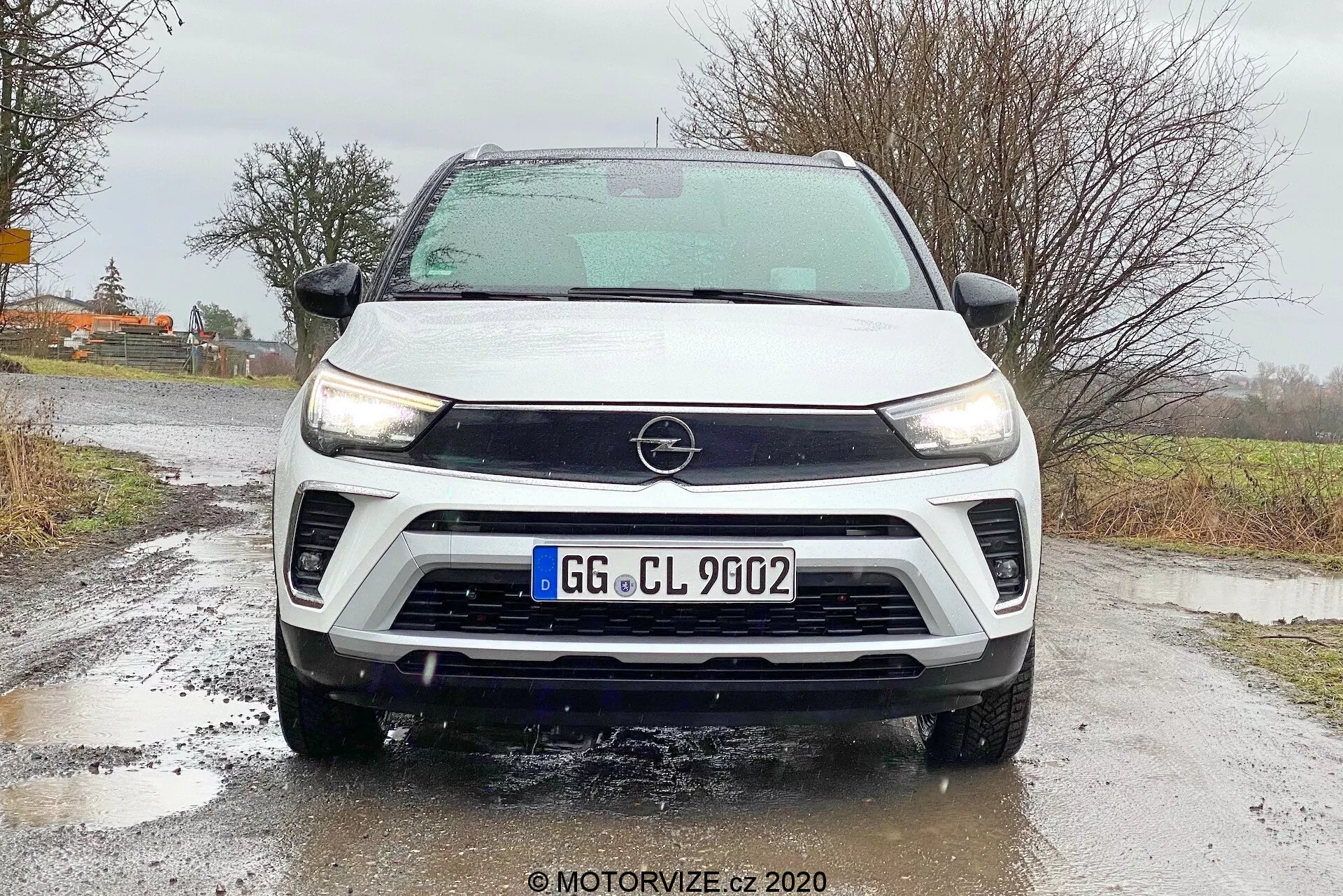 Predný pohľad na Opel Crossland 2021 so zobrazením detailných funkcií, vrátane značky Opel na mriežke, elegantných denných svetiel LED, dvojfarebnej schémy bielej a čiernej farby, predného nárazníka s hmlovkami, širokých nasávaní vzduchu a viditeľnou prednou kamerou indikujúcou pokročilé technológie asistencie vodiča. Auto je stacionárne na mokrom asfalte s viditeľnými kvapkami vody na vozidle, čo zvýrazňuje podmienky riadenia spojené s dažďom.