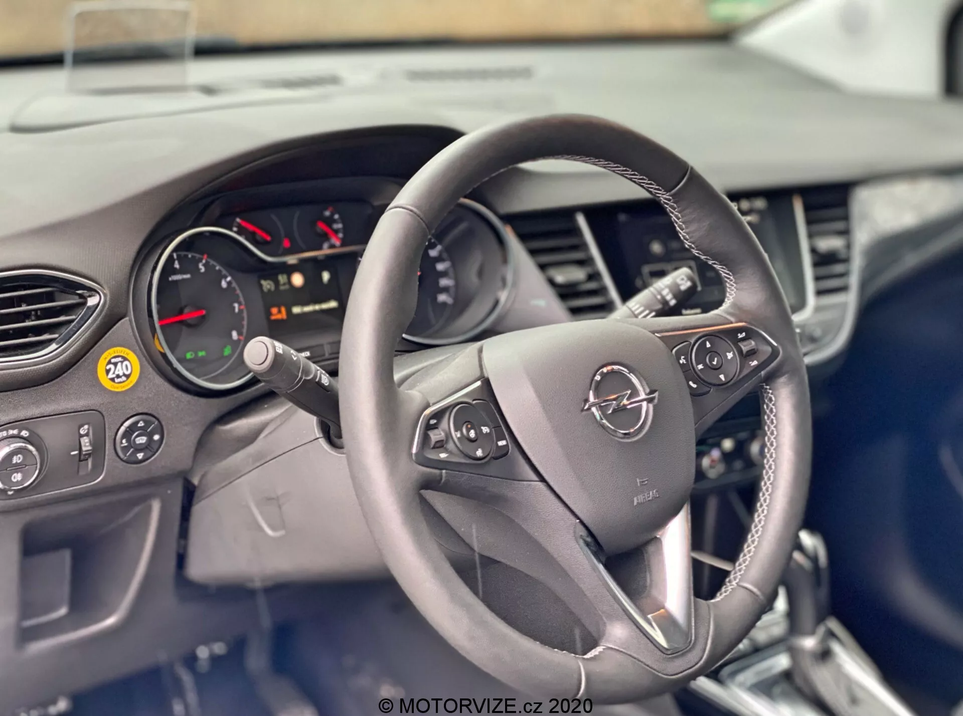Tento obraz ukazuje interiér vozidla Opel Crossland 2021 z pohľadu vodiča, so zameraním na volant a palubnú dosku. Centrálne umiestnený volant má na oboch ramenách ovládacie tlačidlá pre funkcie ako ovládanie audia, hands-free telefon a tempomat. Za volantom je čiastočne viditeľný prístrojový panel s otáčkomerom a rôznymi výstražnými indikátormi. Palubná doska obsahuje ventilačné otvory a čiastočne zakrytý infotainment systém, čo naznačuje moderný a technologicky vybavený interiér.