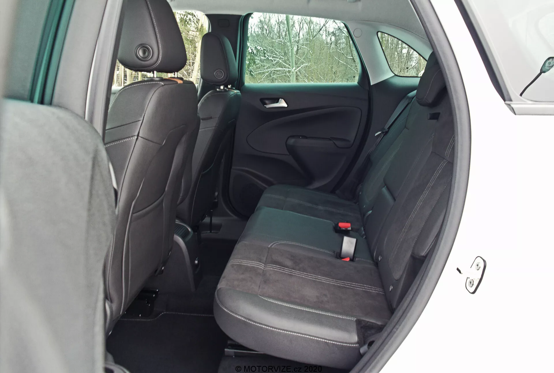 Priestor pre zadných pasažierov Opel Crossland 2021, pohľad z vonkajšieho bočného uhla s otvorenými zadnými dverami. Fotografia, ktorá bola urobená z perspektívy mierne za zadnými dverami, ponúka jasný pohľad do kabíny, ukazujúci dvojfarebné látkové čalúnenie v šedej a čiernej farbe, nastaviteľné hlavové opierky, trojbodový bezpečnostný pás pre stredné sedadlo, ukotvenie ISOFIX pre detské sedačky, priestrannosť, stredovú opierku, úložné vrecko na zadnej strane predného sedadla spolujazdca a panel dverí s rukoväťou, mriežkou reproduktora a ozdobnými prvkami.