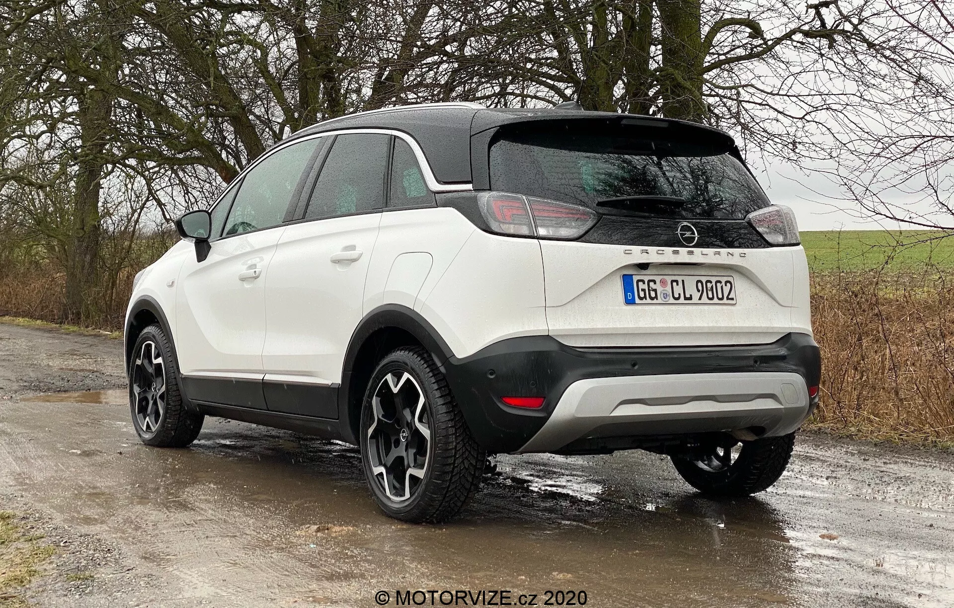 Moderné kompaktné SUV Opel Crossland 2021 v trojštvrťovom zadnom uhle z ľavého zadného pohľadu, zobrazujúce bielu farbu s čiernymi akcentmi, kontrastnou čiernou strechou a bočnými zrkadlami, čierne obklady pozdĺž dolných hrán a oblúkov kolies, horizontálne LED zadné svetlá, odznak výrobcu na zadnej kapote, parkovacie senzory v zadnom nárazníku, štýlové zliatinové kolesá s tmavým povrchom, zaparkované na vidieckej ceste s mokrými povrchmi, ktoré naznačujú nedávny dážď alebo vlhké prostredie.