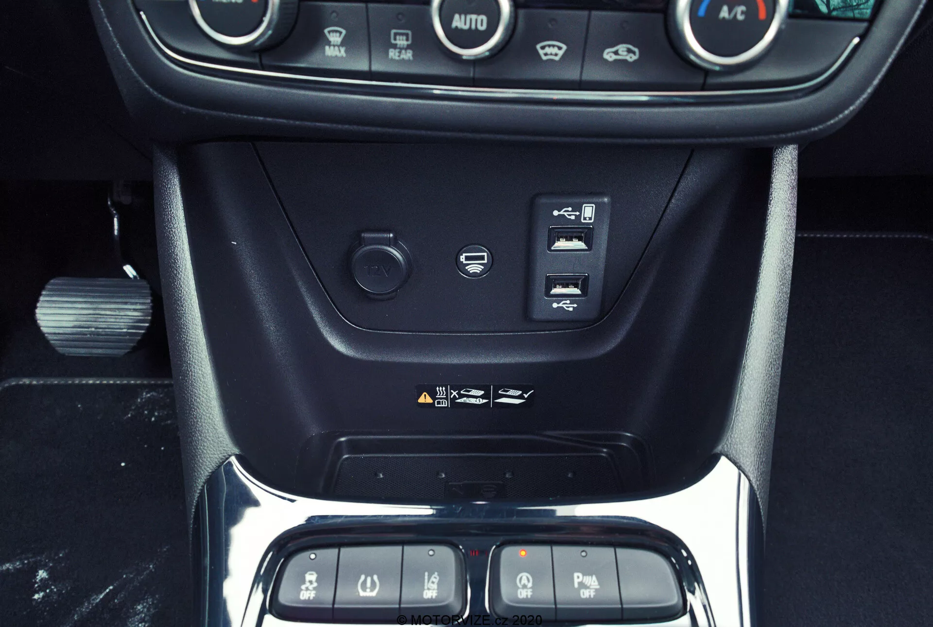 Interiérový záber Opel Crossland 2021 so zameraním na stredovú konzolu, zobrazujúci tlačidlá klimatizačného systému, tlačidlo start/stop motora, tlačidlá asistencie pri riadení, 12-voltovú zásuvku, USB port a časť oblasti radiacej páky s lesklým čiernym panelom orámovaným chrómom, zdôrazňujúc modernú a prémiovú technológiu a dizajn interiéru vozidla.