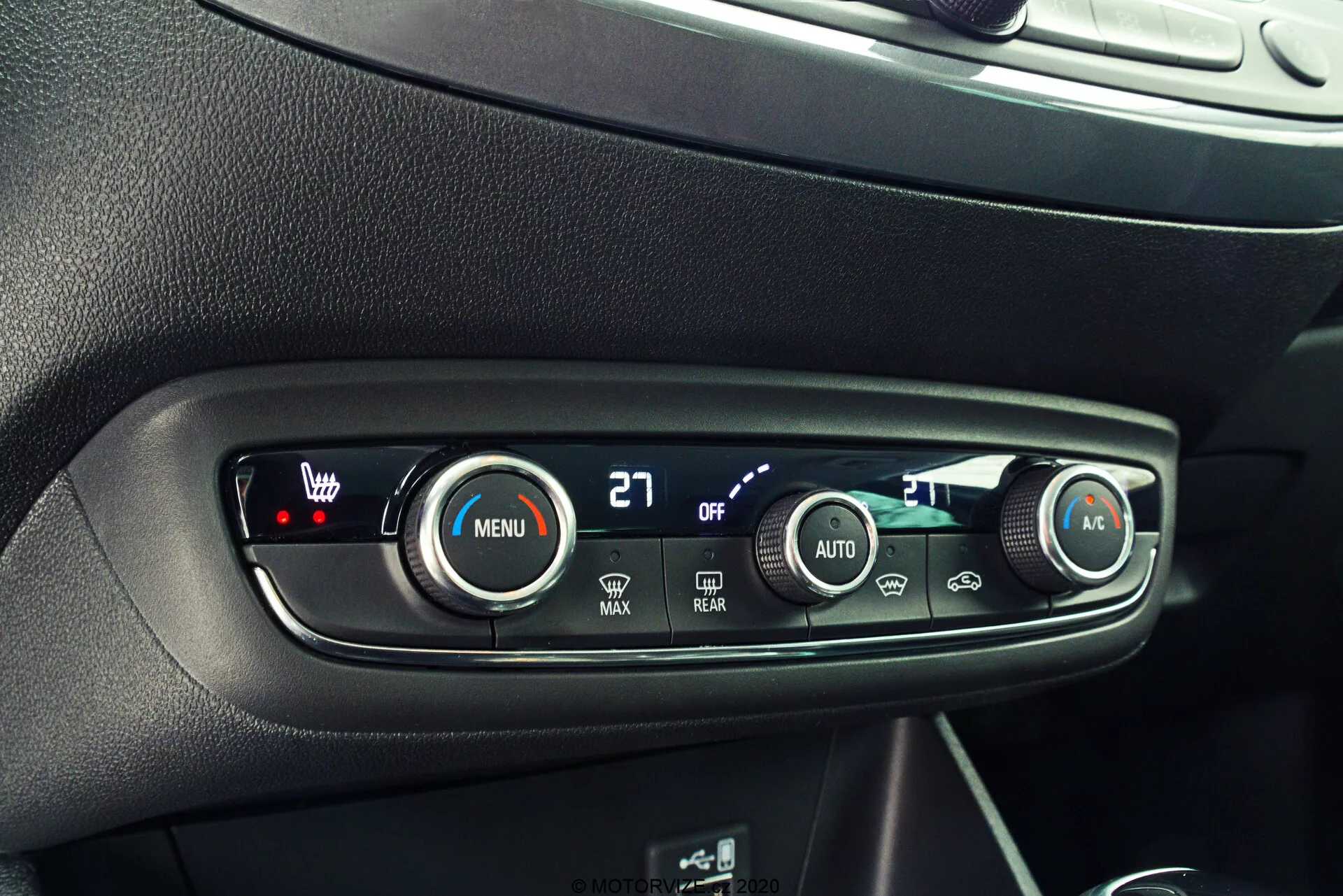 Detailný pohľad na ovládací panel klímy v interiéri auta, ktorý sa vyznačuje elegantným, moderným dizajnom s čiernymi povrchmi, striebornými akcentmi a jasnými bielymi popiskami. Čelná, mierne sklonená perspektíva zdôrazňuje ovládacie tlačidlá a gombíky. Ľavý gombík s červenou LED a zobrazenou hodnotou '27' ovláda teplotu pre vodiča. Tlačidlo 'MENU' s ikonou ventilátora umožňuje prispôsobenie nastavení ventilátora. Menší tlačidlo 'OFF' pravdepodobne vypína systém. Tlačidlá ventilátora 'MAX', odmrazovanie 'REAR' a tlačidlo 'AUTO' pre automatickú klimatizáciu, a pravý gombík s modrou LED pre teplotu spolujazdca dotvárajú pokročilý a funkčný dizajn.