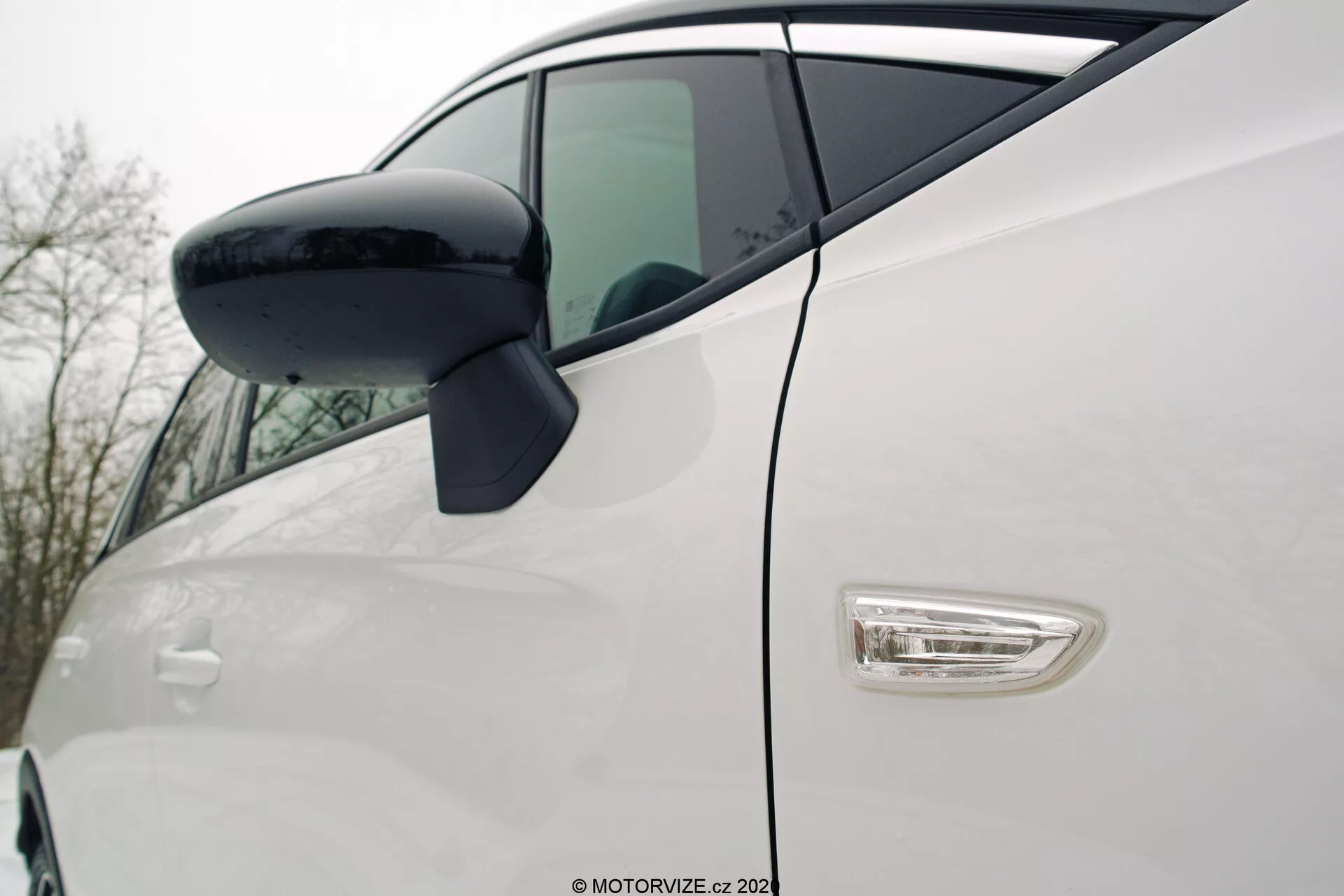 Detailný pohľad z uhla na biely Opel Crossland 2021 z pohľadu na predný štvrtpanel a dvere vodiča. Zahŕňa čierne bočné zrkadlo umiestnené na dverách, elegantné chrómované kľučky s možným bezkľúčovým prístupom, aerodynamický LED bočný indikátor, lesklú bielu farbu a zakrivené línie. Moderný dizajn vozidla zdôrazňujúci technologické a estetické detaily.