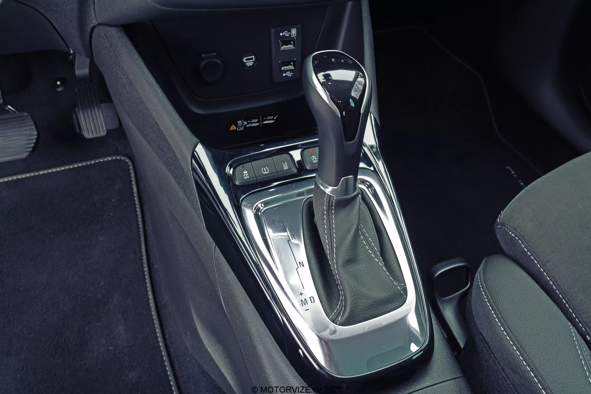 Detailný pohľad zhora na stredovú konzolu Opel Crossland 2021 s automatickou radiacou pákou s koženou rukoväťou, chrómovými a klavírne čiernymi prvkami, elektronickou parkovacou brzdou a možným úložným priestorom alebo panelom s konektormi, obklopený čiernym koženým čalúnením s kontrastným šitím.